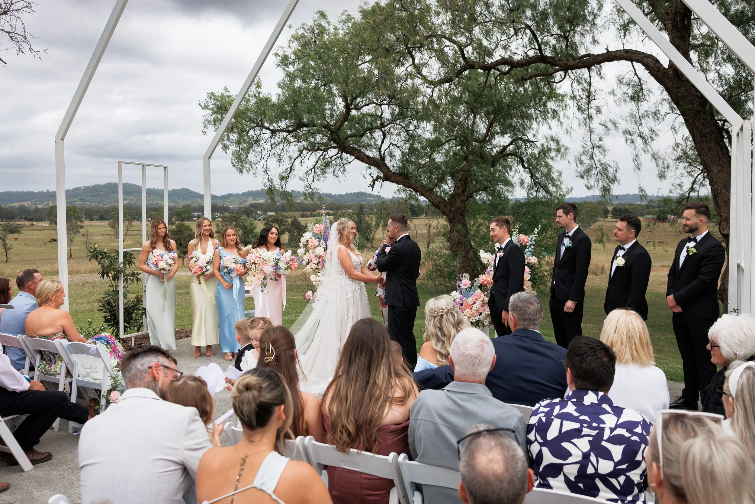 A wedding ceremony taking place outdoors, with the bride and groom holding hands and smiling at each other, surrounded by the wedding party and guests, under a large tree.