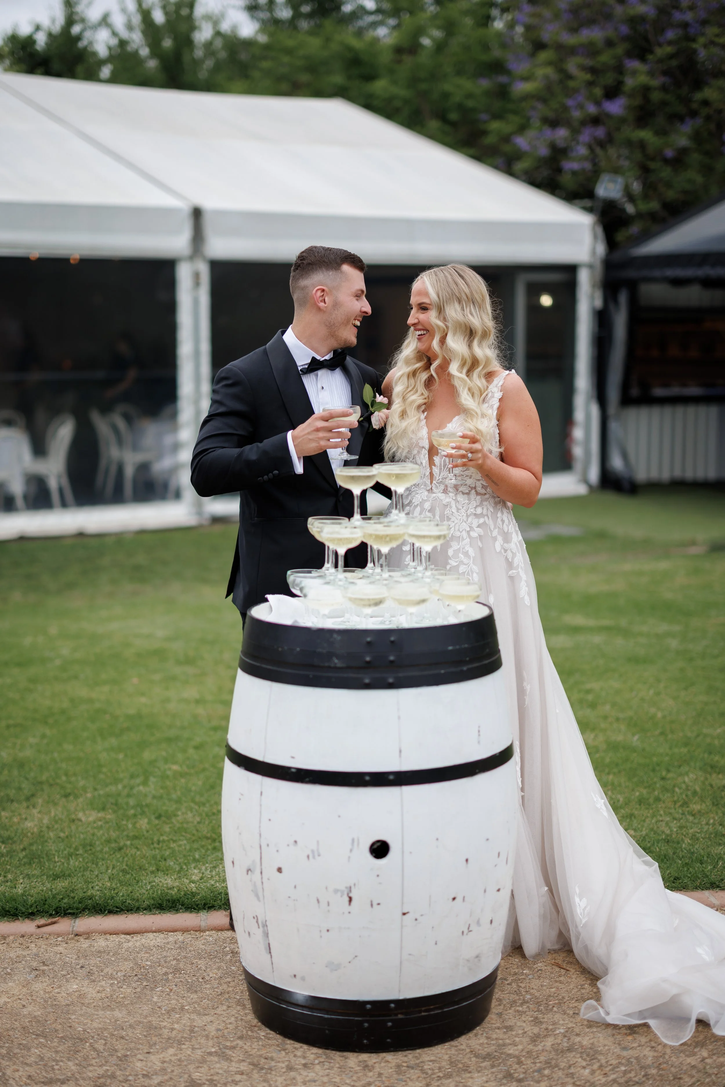 Bride and groom celebrating at wedding reception outdoors, raising glasses near champagne display on a barrel table.