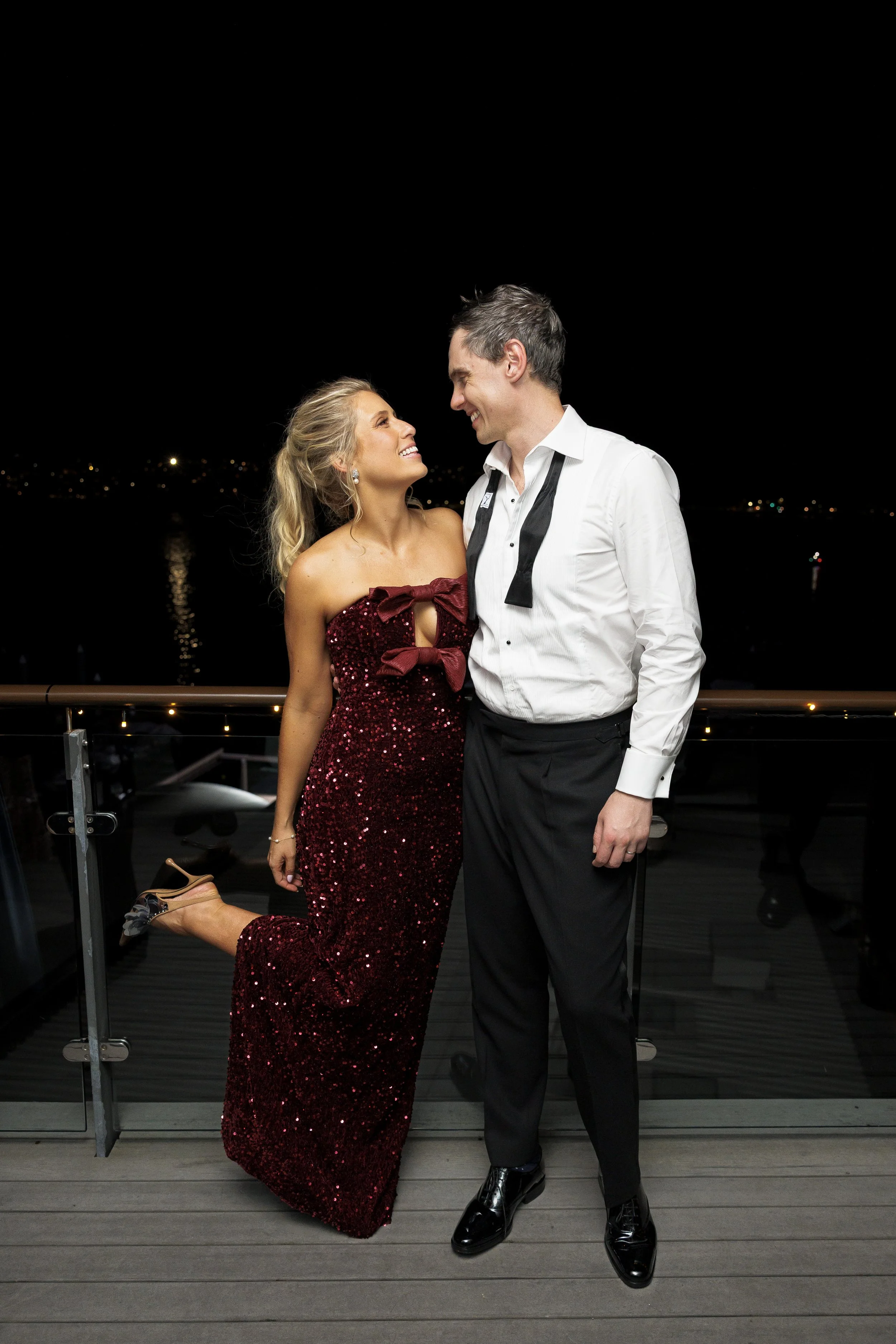 A couple dressed in formal wear, standing on a balcony at night, smiling and looking into each other's eyes. The woman is in a strapless, sequined red gown with a bow on the chest, and one of her legs is raised. The man is in a white dress shirt with