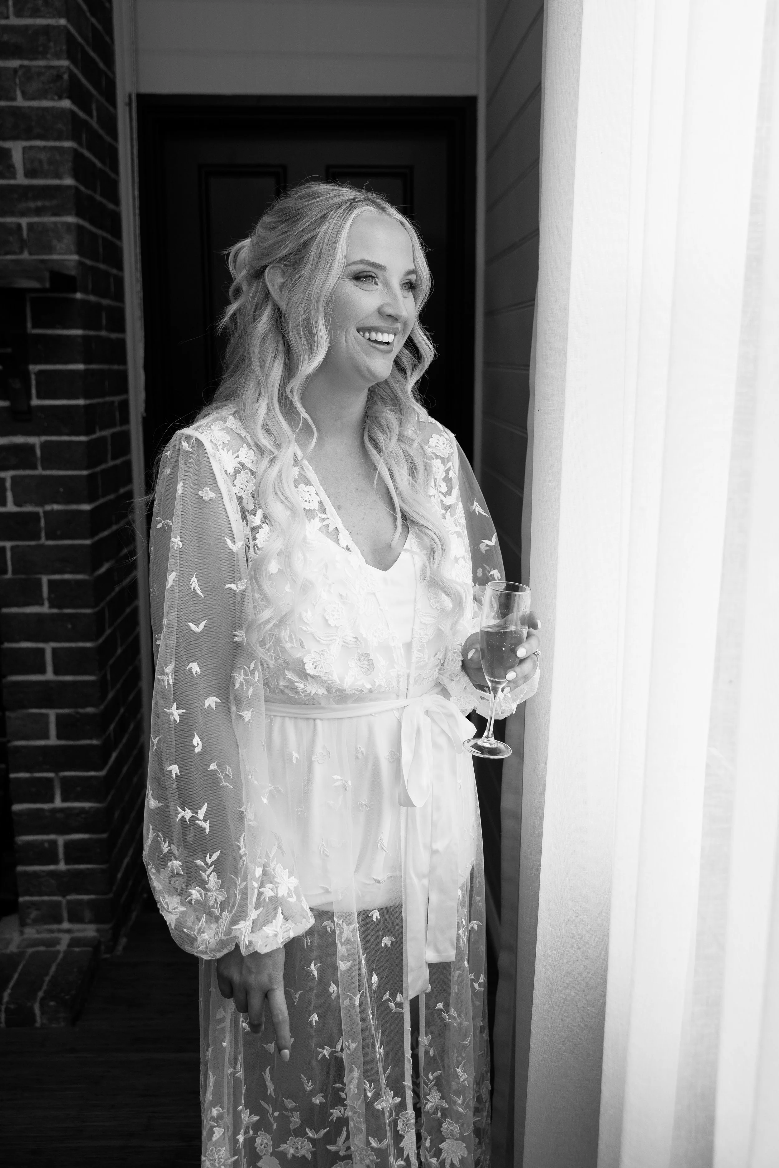 A woman dressed in a robe with floral embroidery, smiling and holding a glass of champagne in front of a window with sheer curtains.