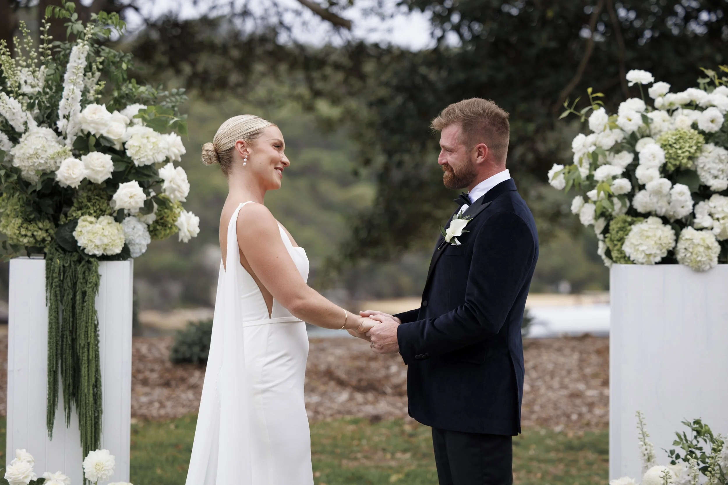 A bride and groom holding hands during their wedding ceremony outdoors, surrounded by large white floral arrangements and trees.