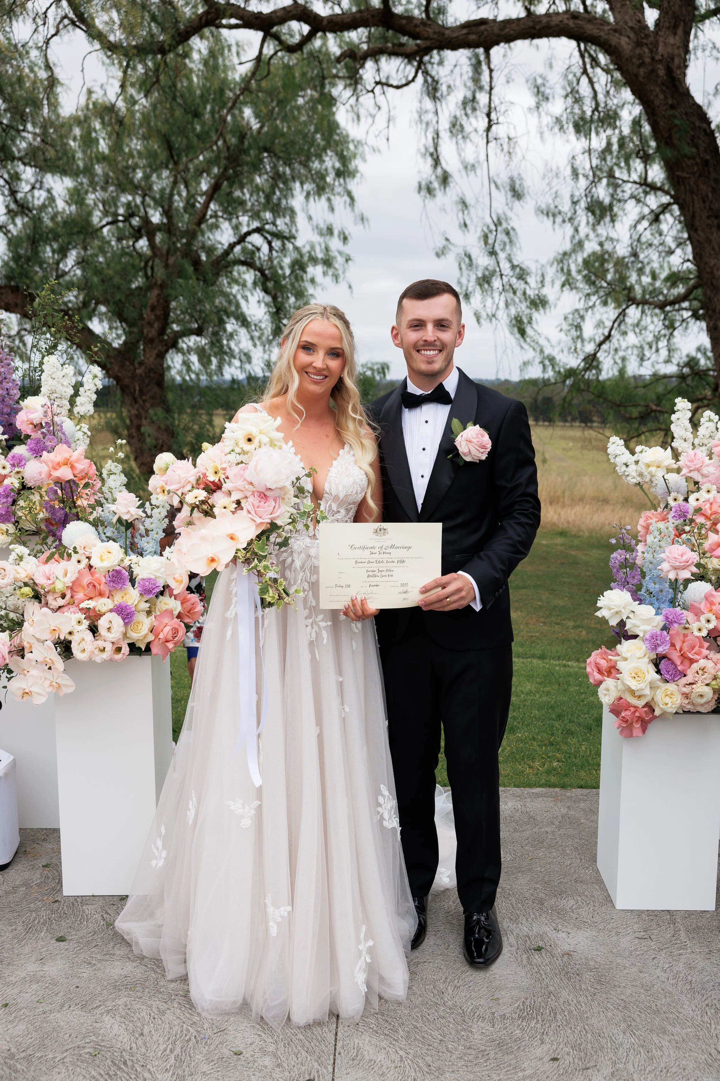 A bride and groom standing outdoors during their wedding, smiling and holding a marriage certificate, surrounded by large floral arrangements with pink, white, and purple flowers.