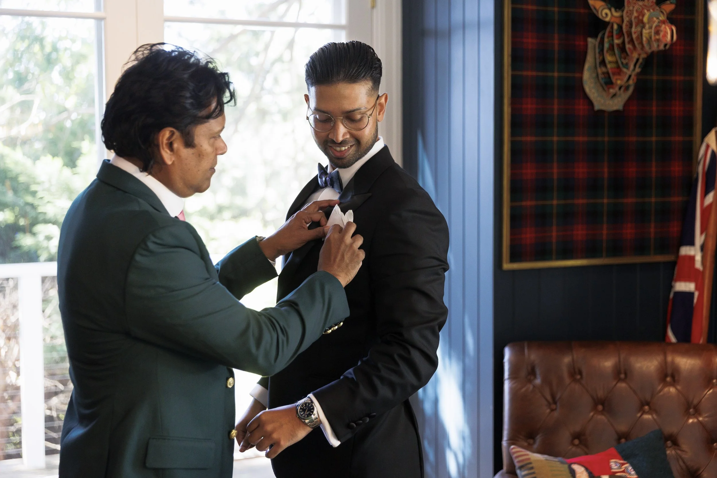 A man in a tuxedo with a bow tie smiling while an older man in a green blazer pins a badge or pin onto his lapel inside a room with a window and a Union Jack flag.