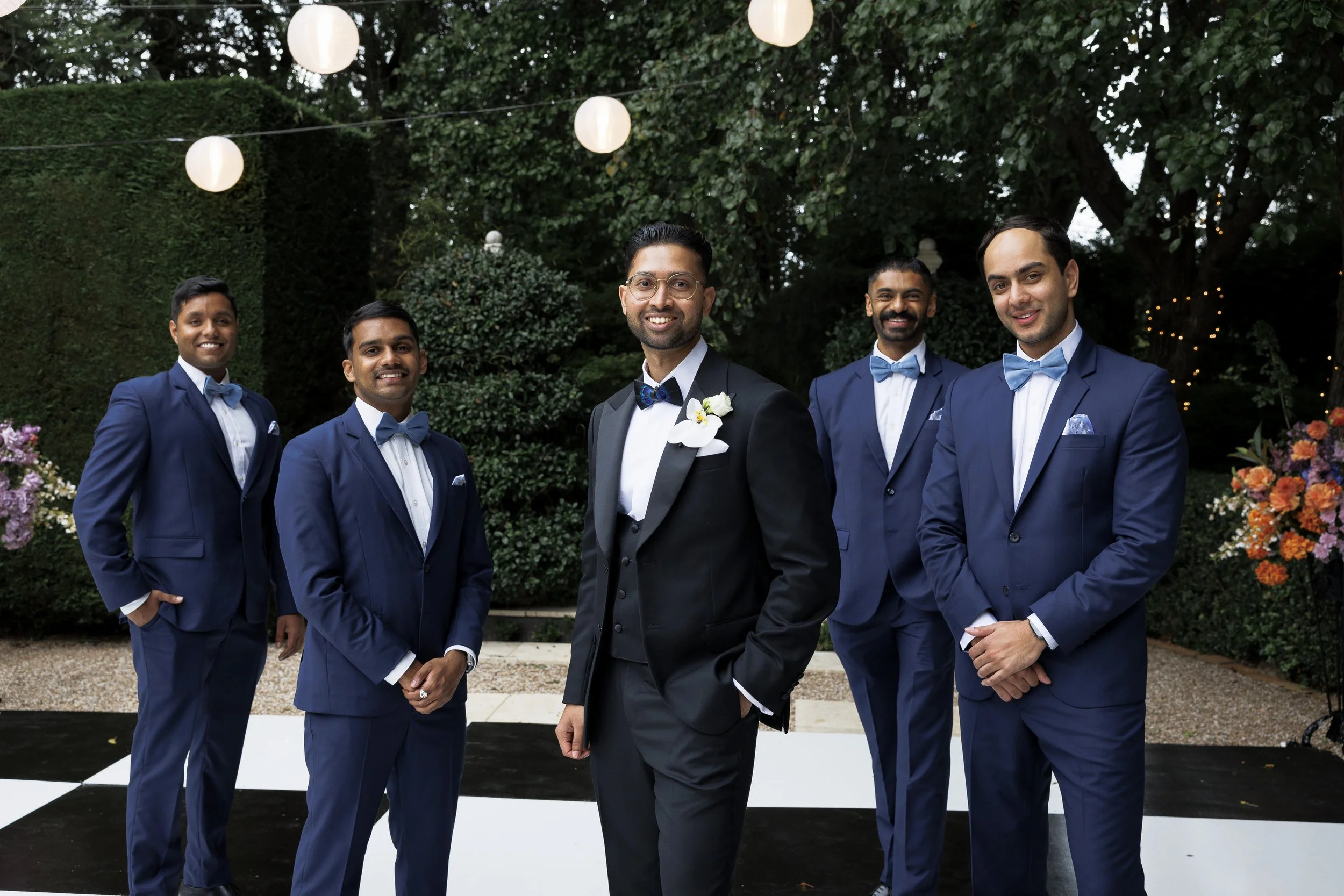 A groom in a black tuxedo with a white shirt and a blue bow tie, standing with five groomsmen in navy suits with light blue bow ties, outdoors under string lights, with greenery and flowers in the background.