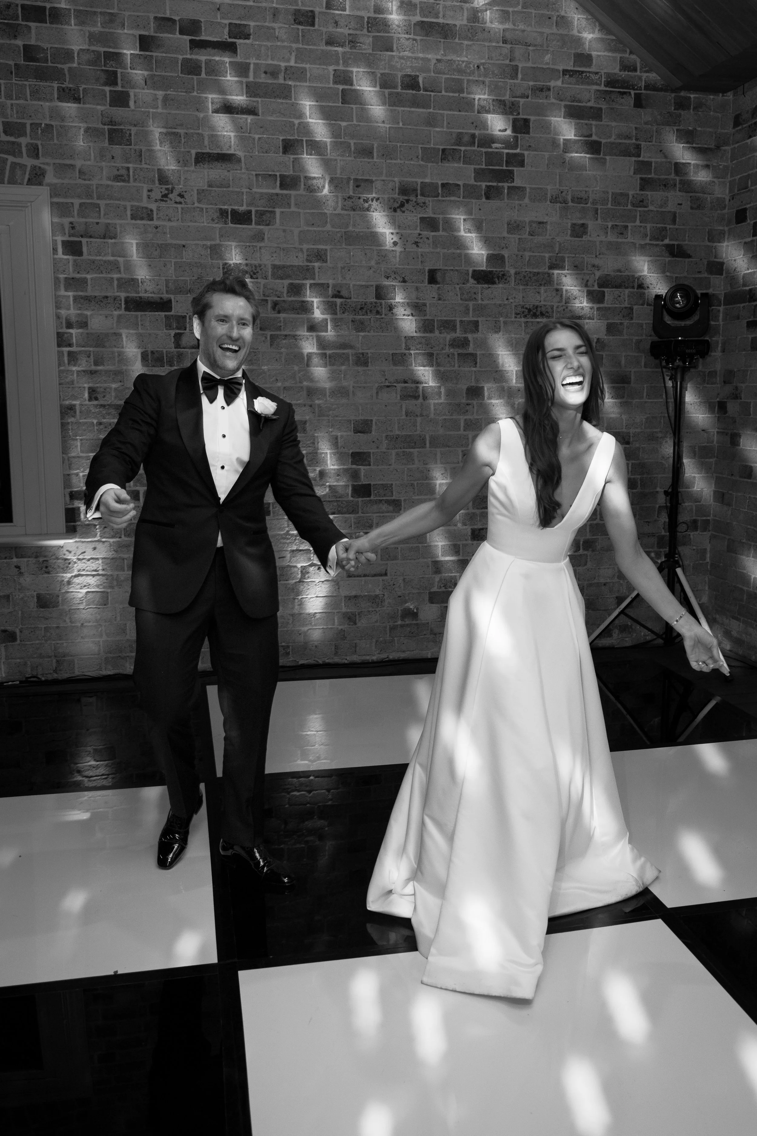 A black and white photo of a newlywed couple dancing at their wedding reception, smiling and holding hands in front of a brick wall with dance floor lighting.