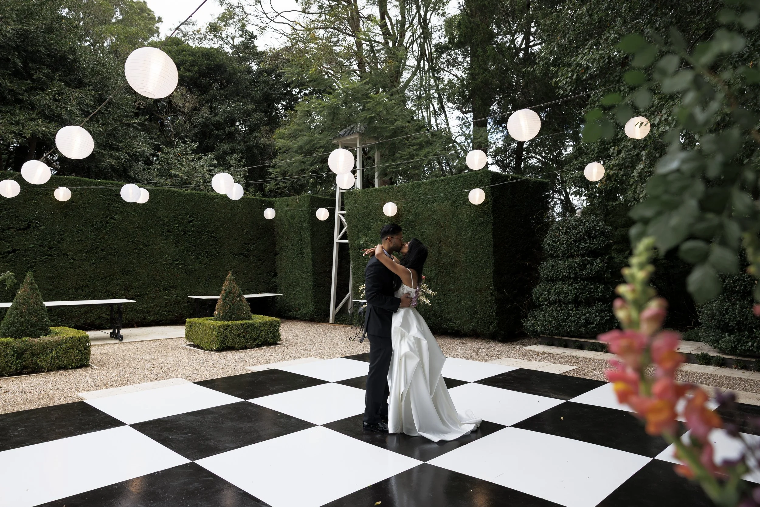A newlywed couple sharing a kiss on a black and white checkered dance floor outdoors, surrounded by greenery and decorated with hanging white lanterns.