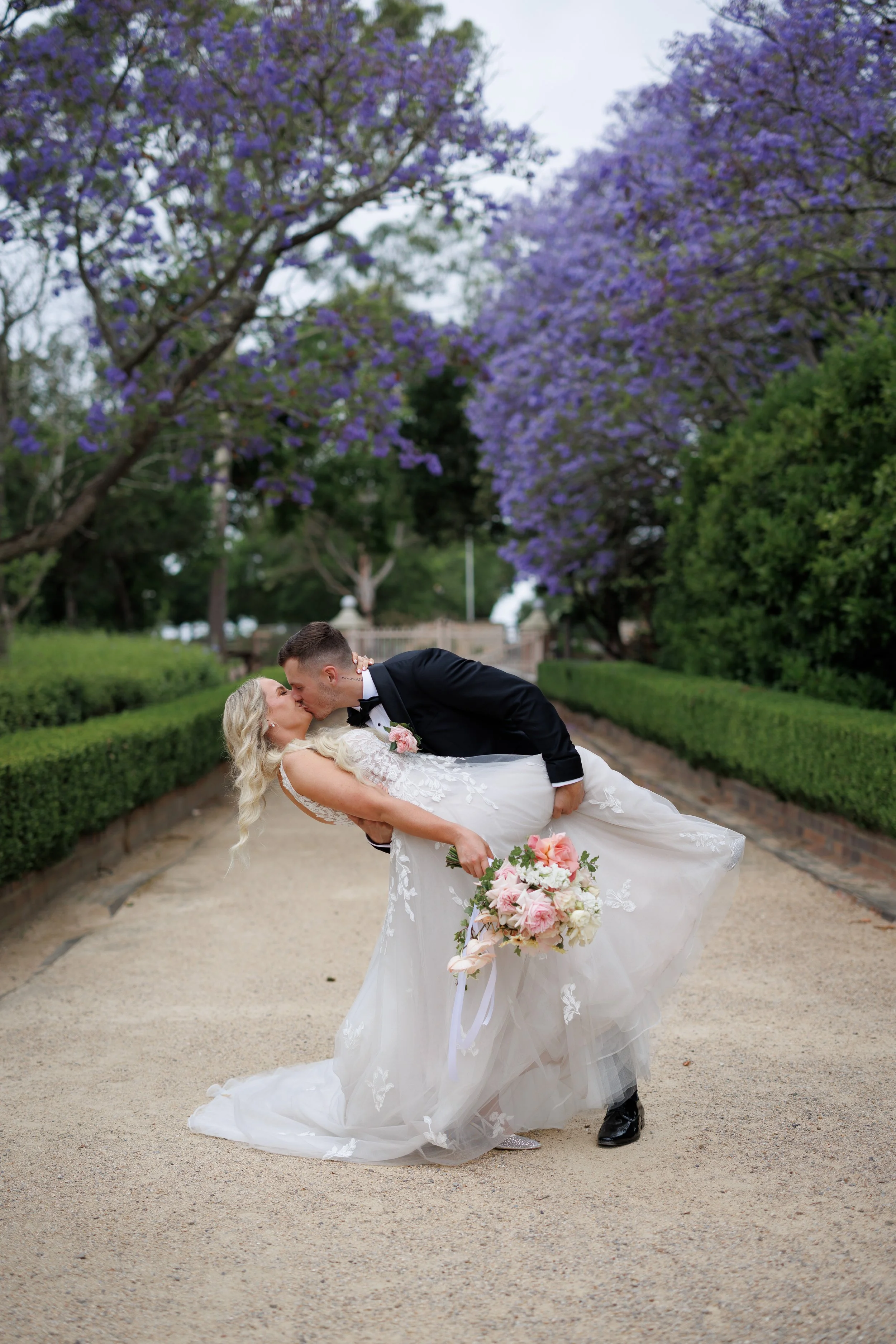 A bride and groom sharing a kiss on a garden pathway during their wedding, with purple flowering trees in the background.