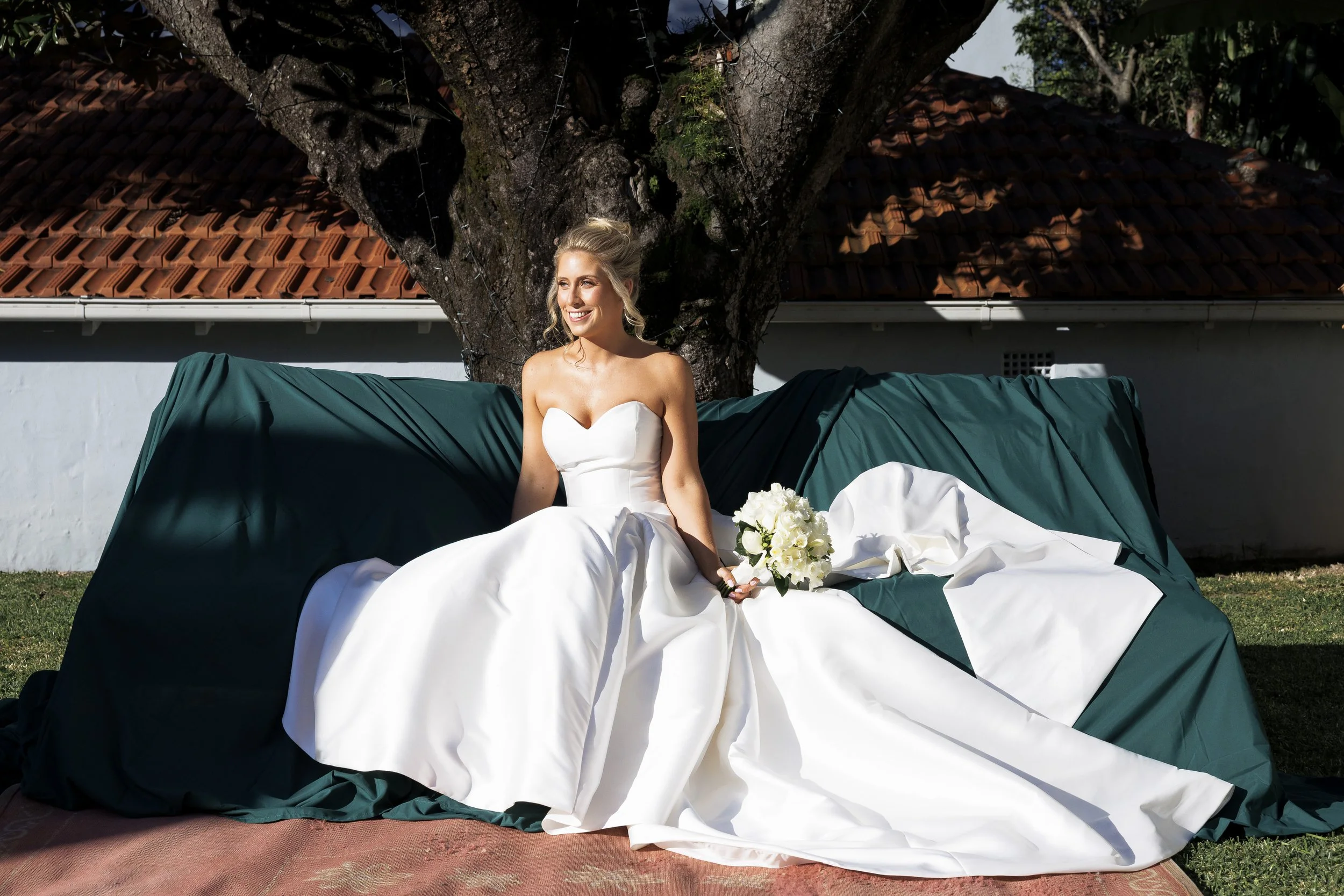 A bride in a strapless white wedding gown holding a bouquet of white flowers, sitting outdoors on a green-covered bench in front of a large tree with a red-tiled roof house in the background.