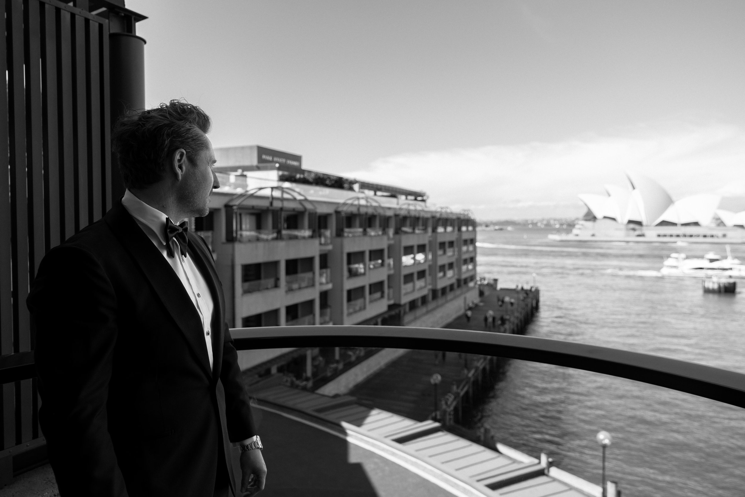 Man in tuxedo looking out from a balcony over a city waterfront with buildings and the Sydney Opera House in the background.