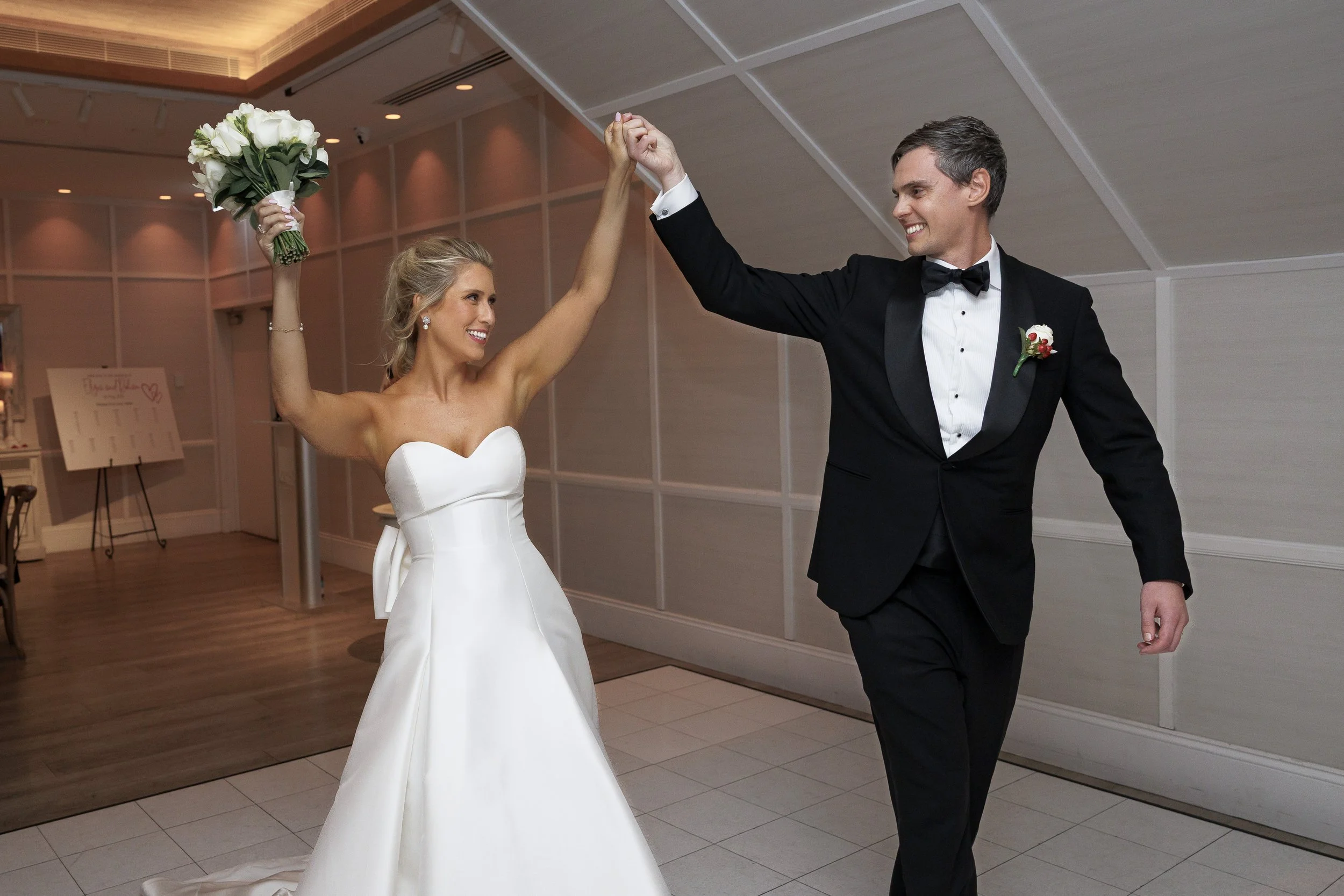 A bride and groom happily dance together at their wedding reception, holding hands up in celebration, with the bride smiling and holding a bouquet of white roses.