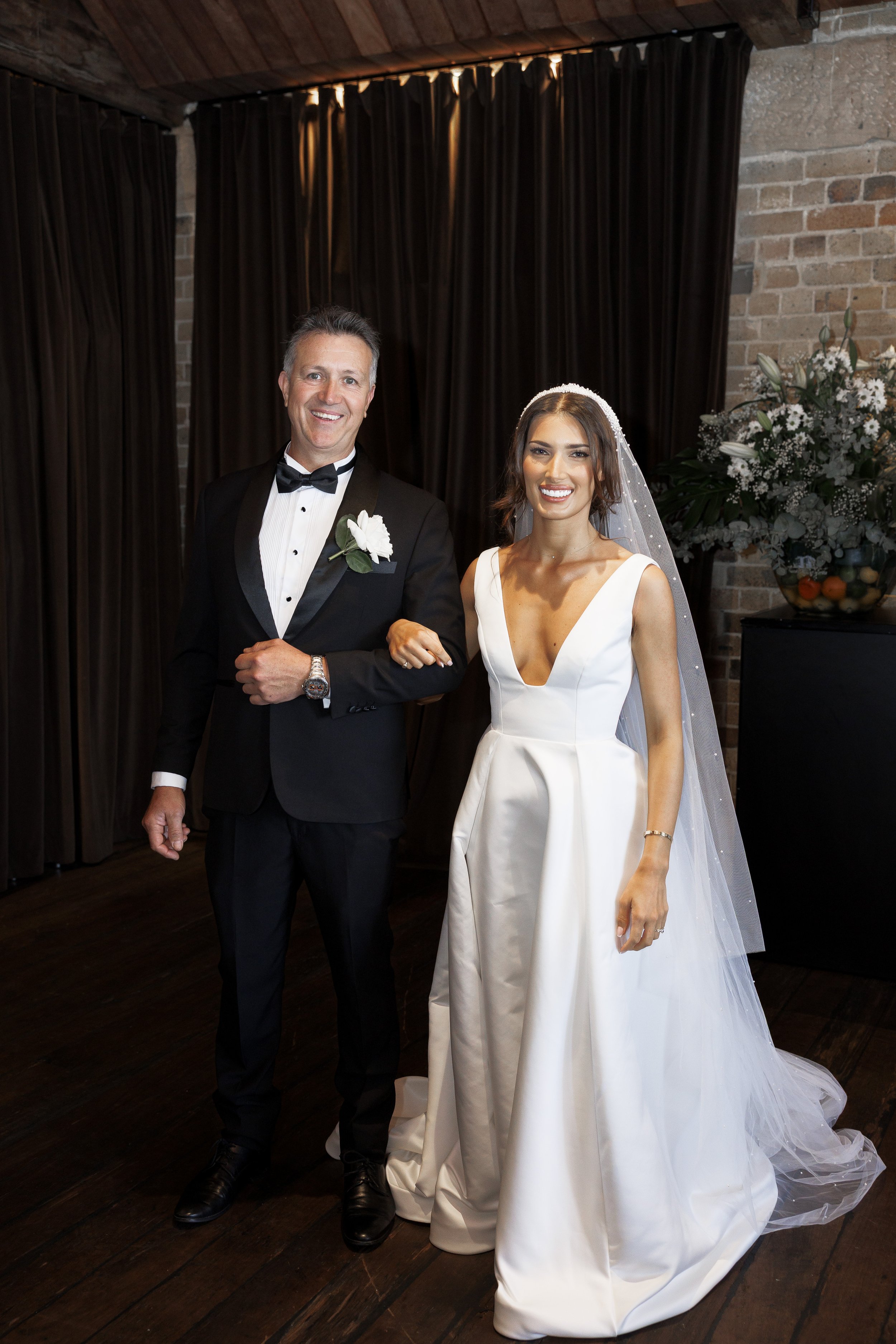 A bride in a white wedding gown standing next to a groom in a black tuxedo, both smiling at the camera, indoors with dark curtains and brick walls.