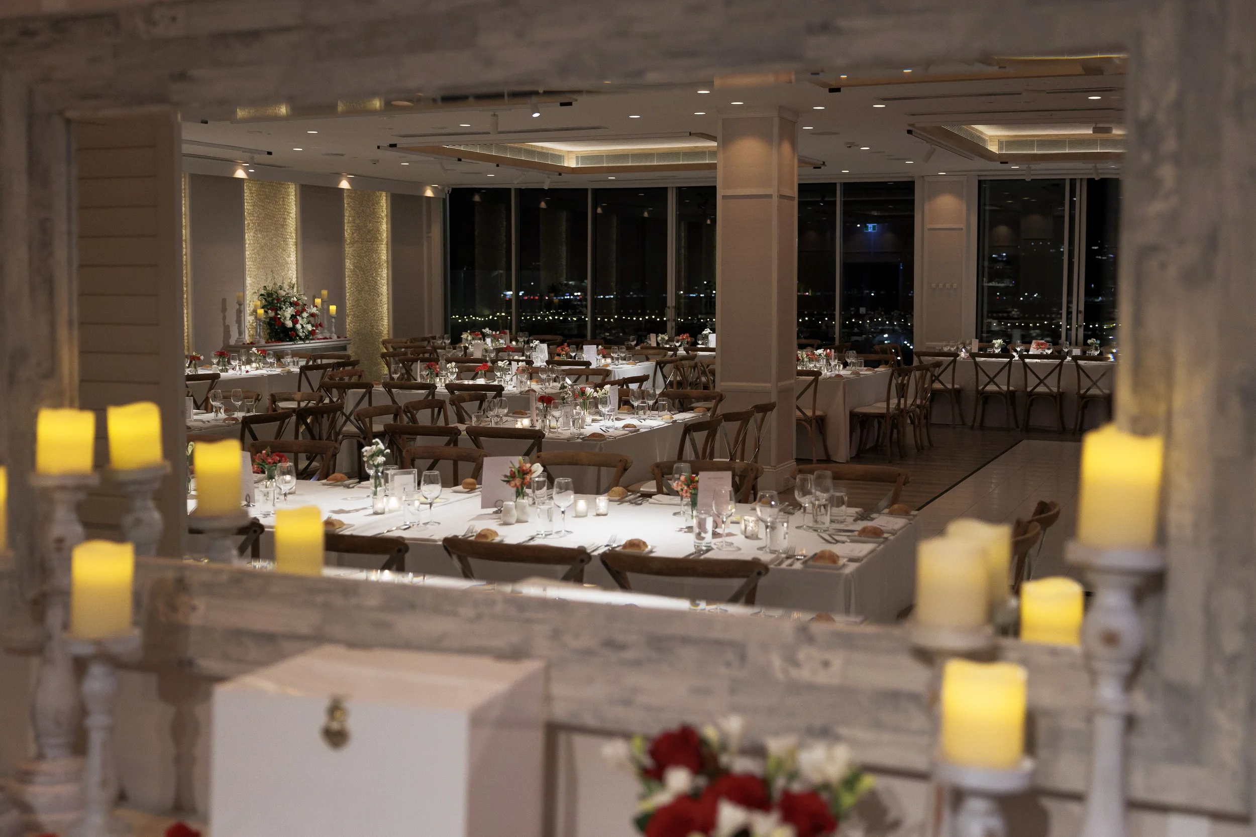 Elegant banquet hall decorated for a formal event with round and rectangular tables, white tablecloths, floral centerpieces, and candles, viewed through a window frame, with city lights visible outside.
