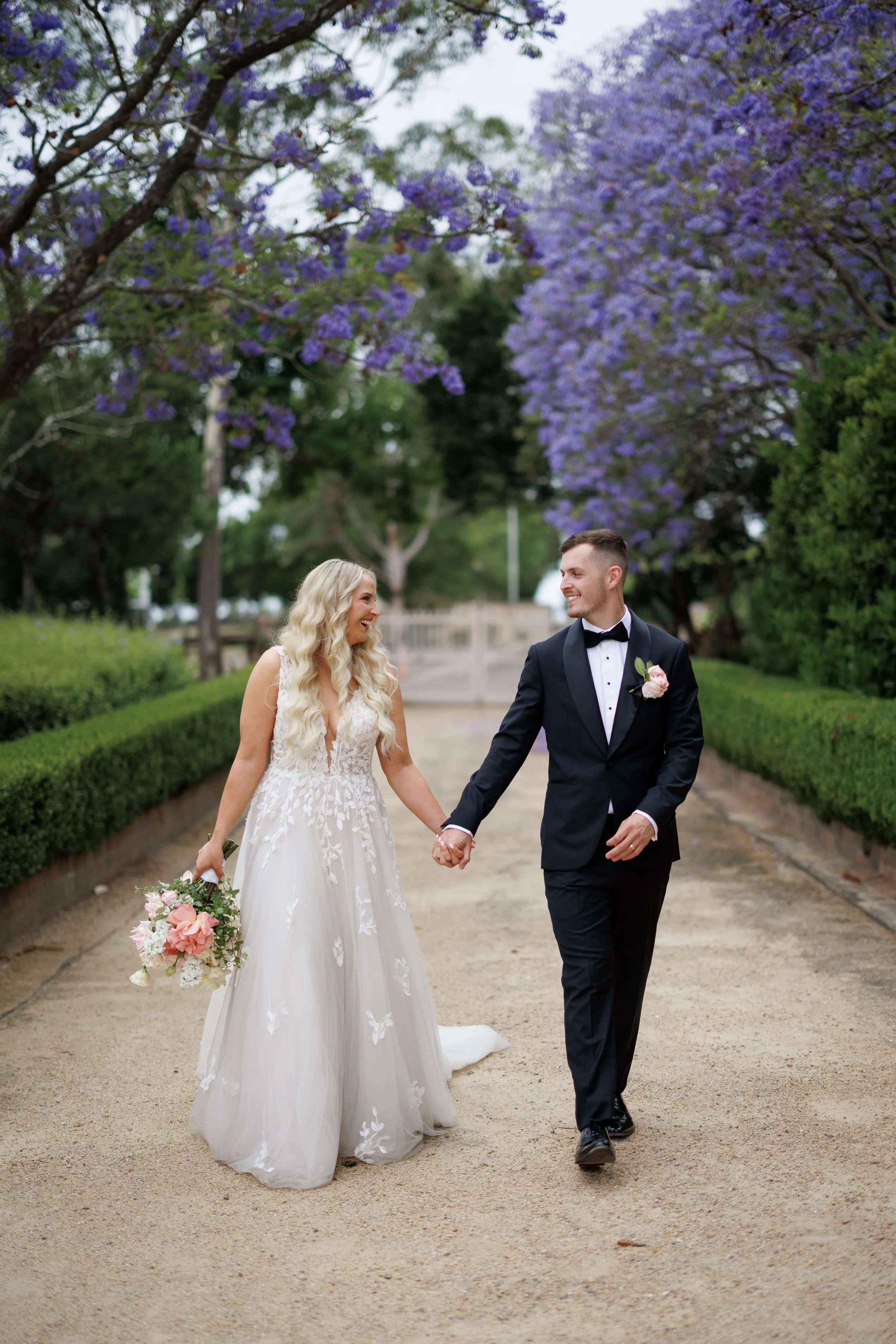 Bride and groom walking hand in hand on a garden path, surrounded by blooming purple jacaranda trees.