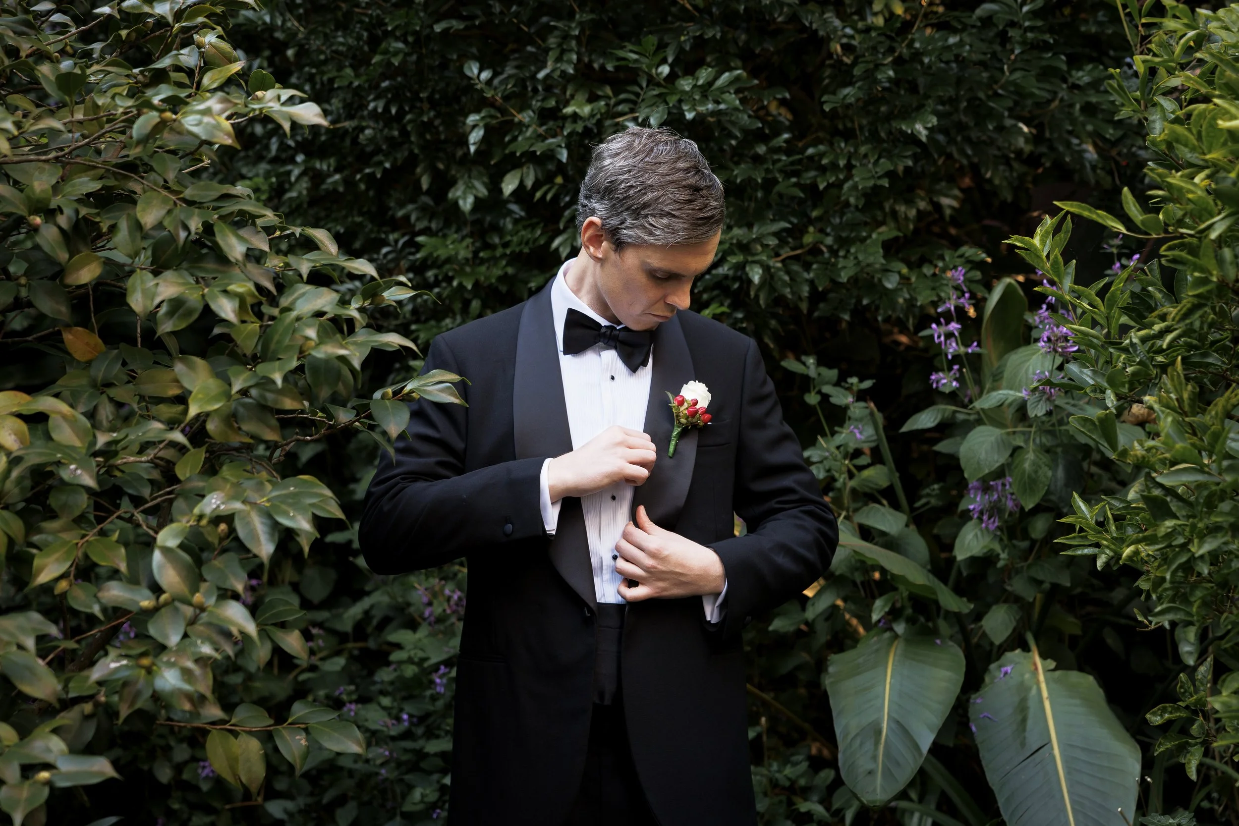 A man in a black tuxedo with a bow tie and white shirt adjusting a boutonniere on his lapel, standing among green foliage and purple flowers.