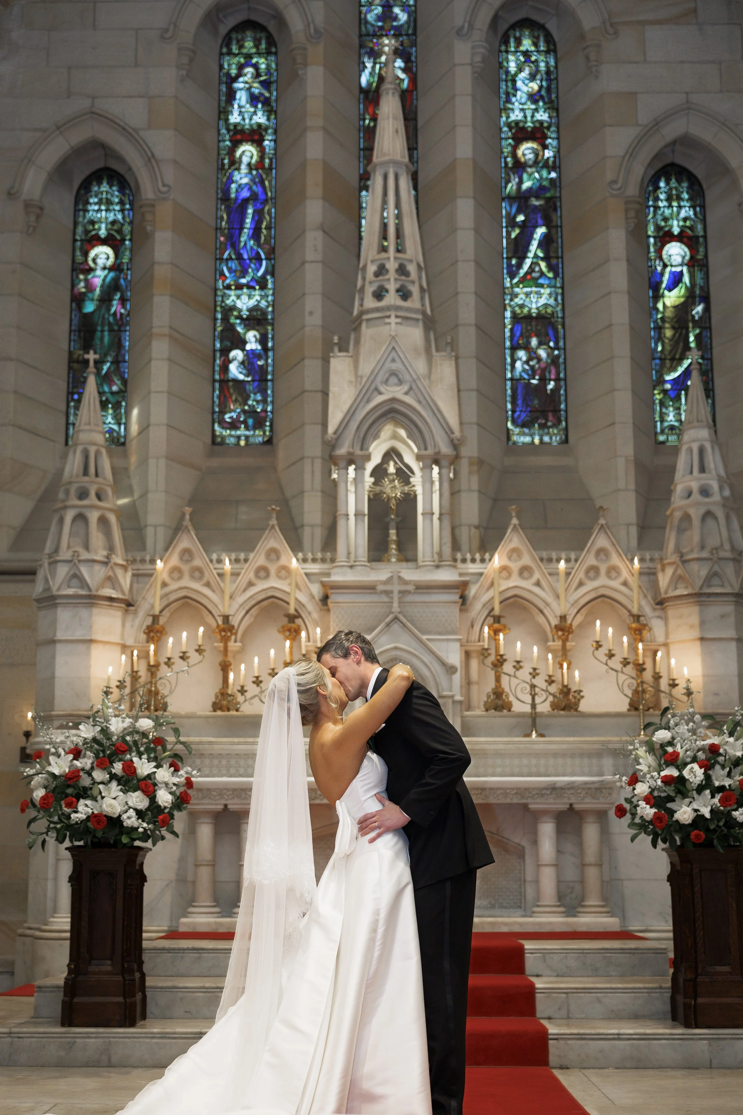 A bride and groom kissing at their wedding ceremony inside a church with stained glass windows and floral arrangements.