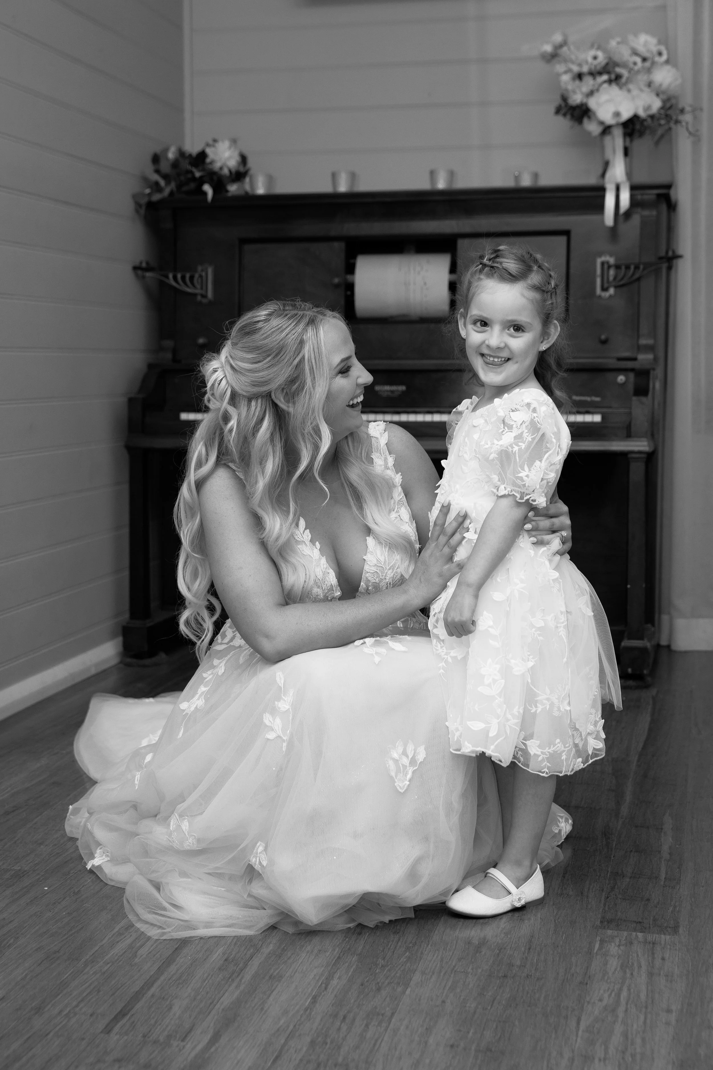 A woman and a girl sharing a joyful moment indoors, with the woman sitting on the floor and the girl standing. Both are dressed in light-colored dresses, and there is a piano and floral arrangements in the background.