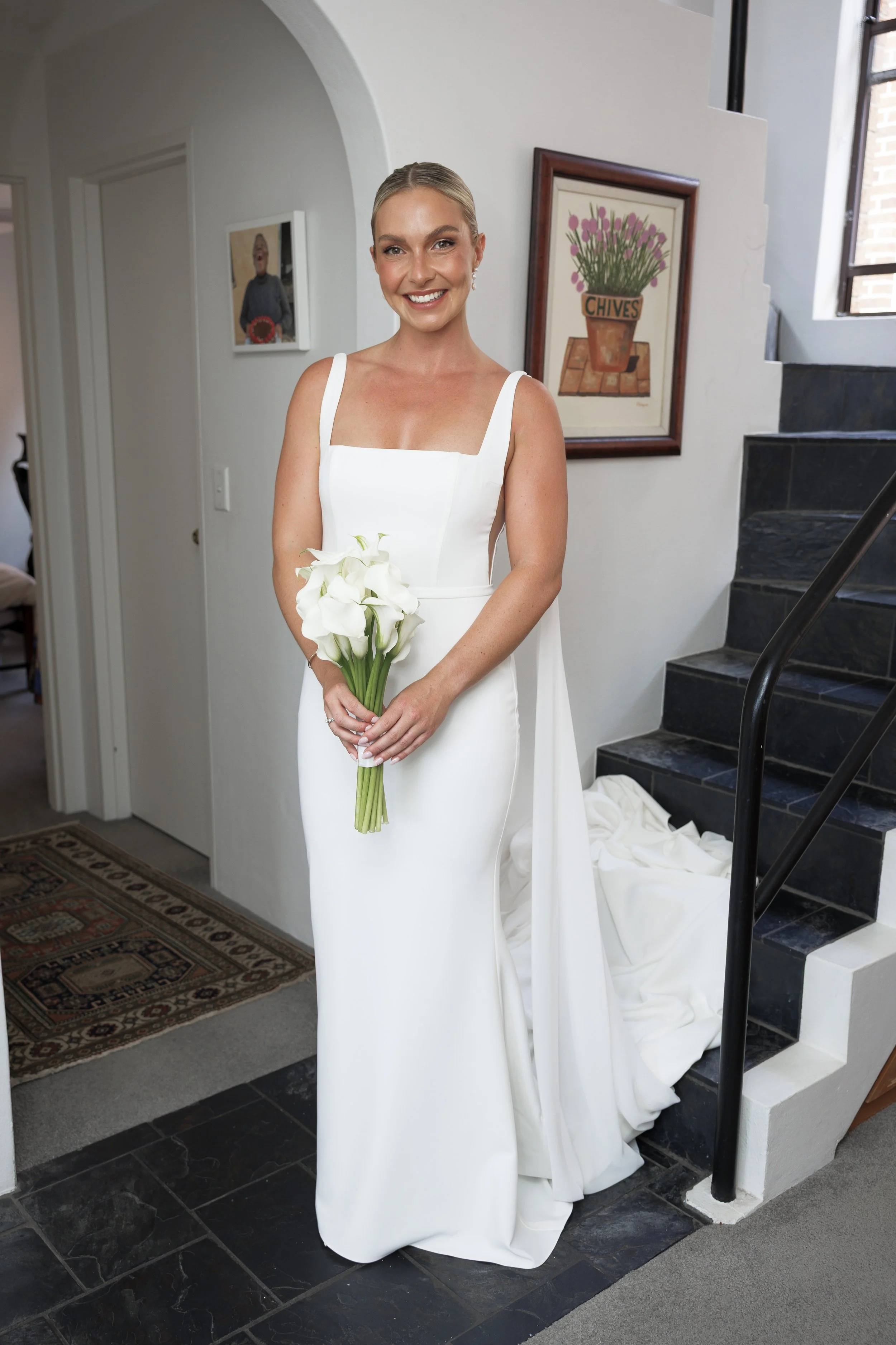 A woman in a white wedding dress holding a bouquet of white calla lilies smiling in a home interior with staircase and framed art on the wall.