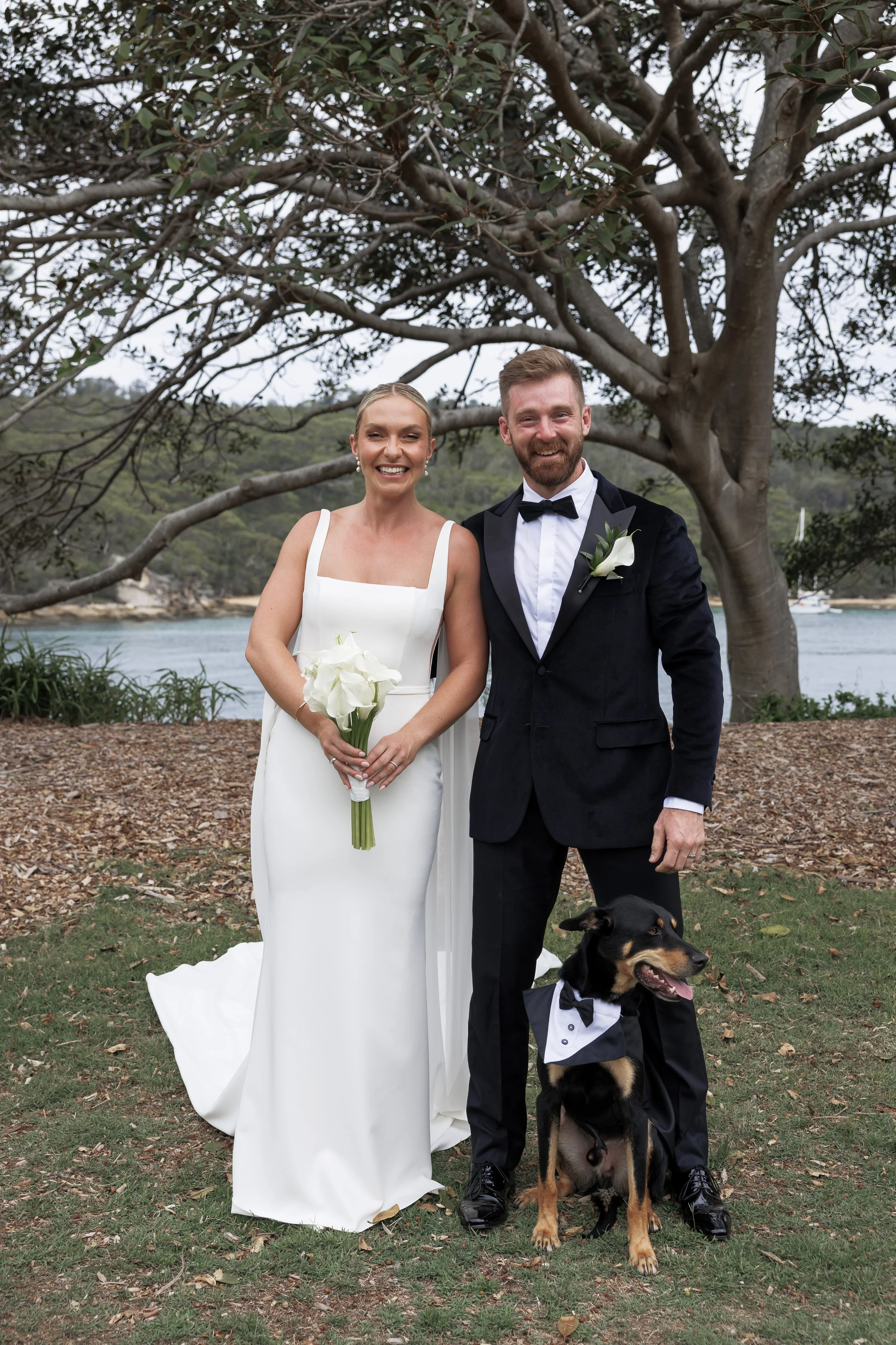 A bride and groom pose outdoors on their wedding day with a black and brown dog wearing a tuxedo. The bride is in a white gown holding a bouquet of white calla lilies, and the groom is in a black tuxedo with a bow tie and boutonniere. They are standi