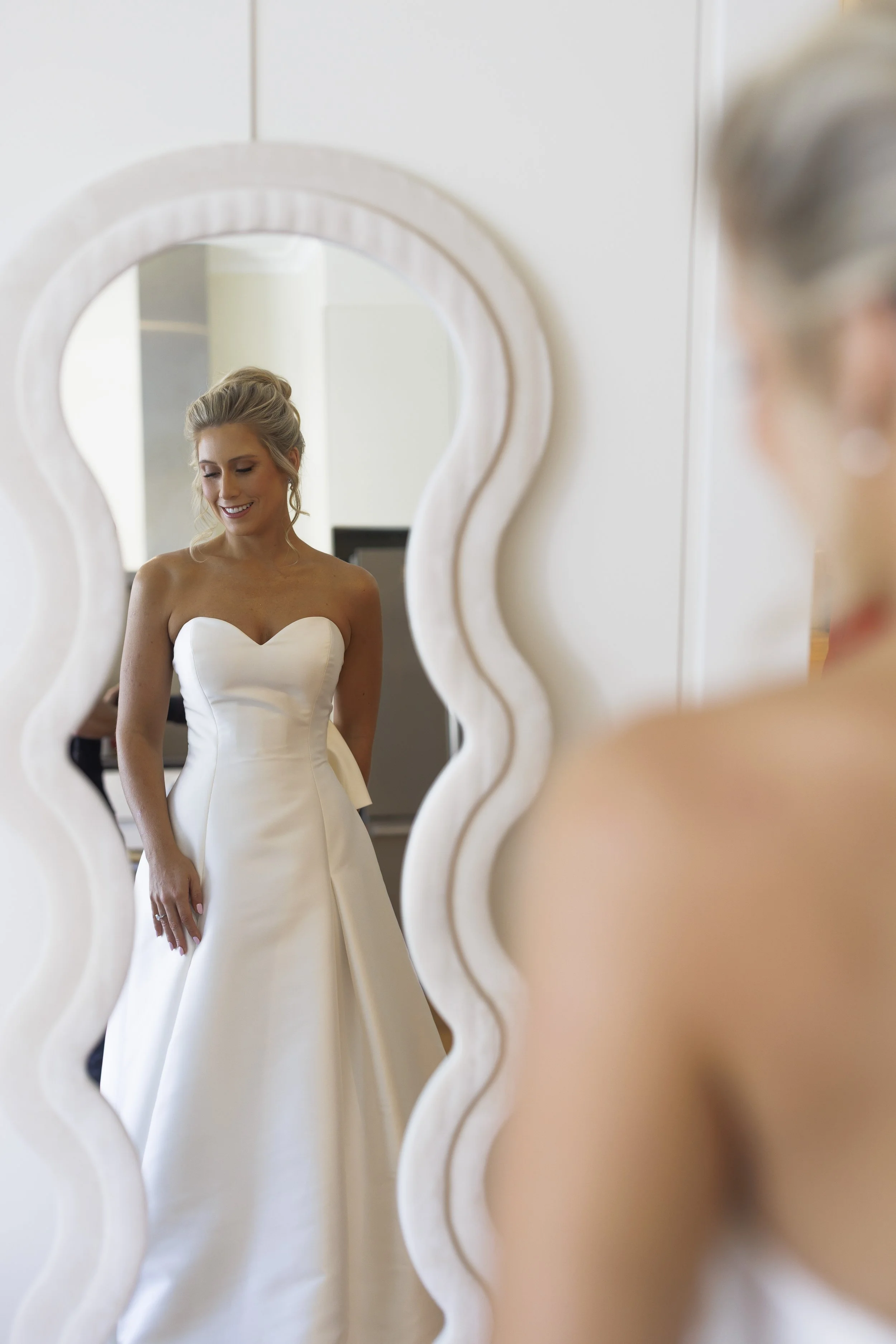 A woman in a strapless white wedding dress smiling as she looks at herself in a mirror with a scalloped white frame.