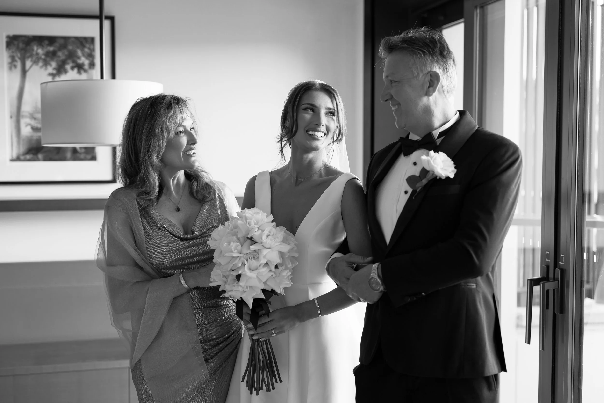 A bride holding a bouquet of flowers smiles at a groom in a tuxedo as his mother looks on, in a room with large windows and framed artwork in the background.