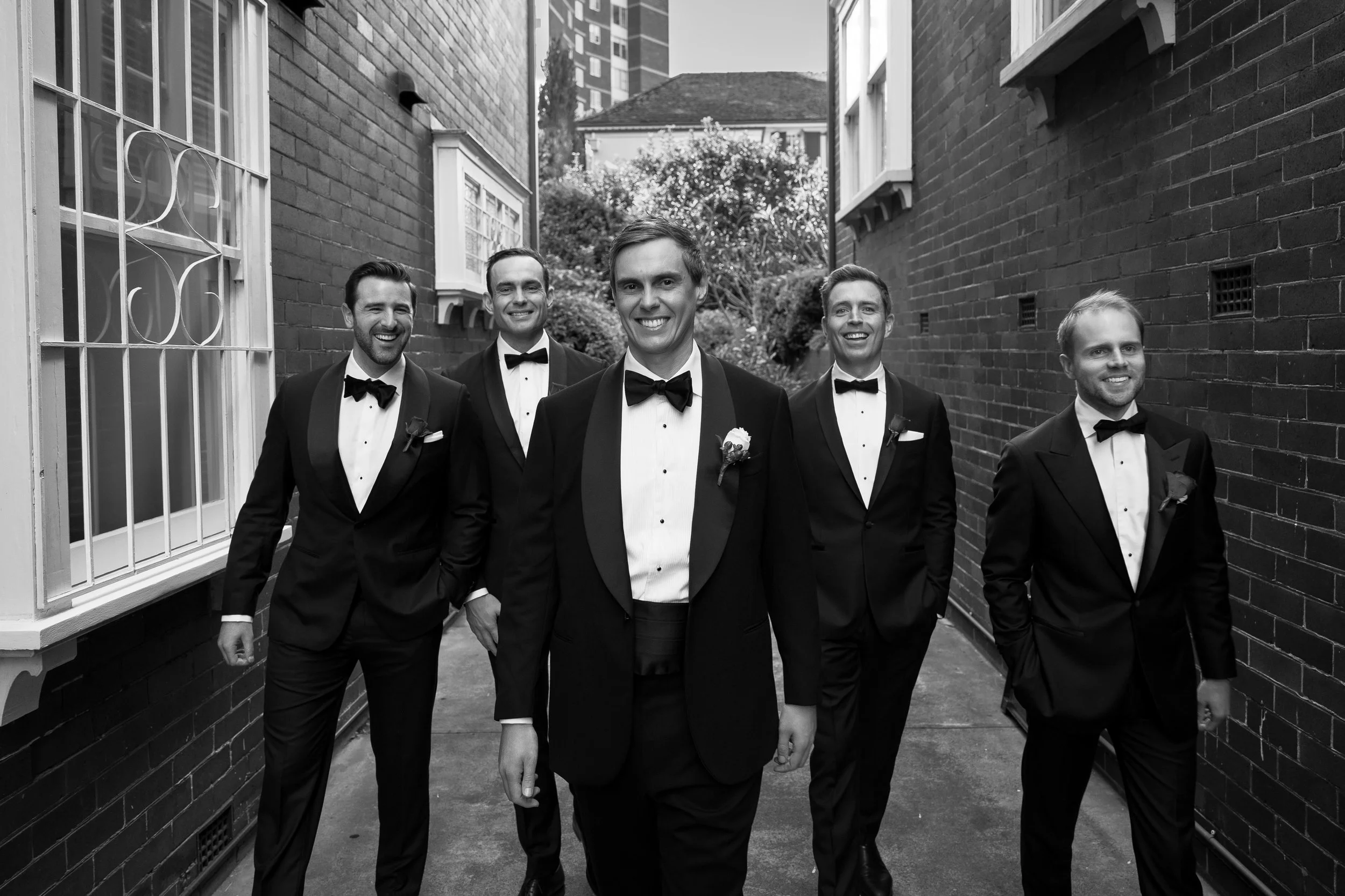 Group of five men in tuxedos walking outdoors between brick buildings, smiling.