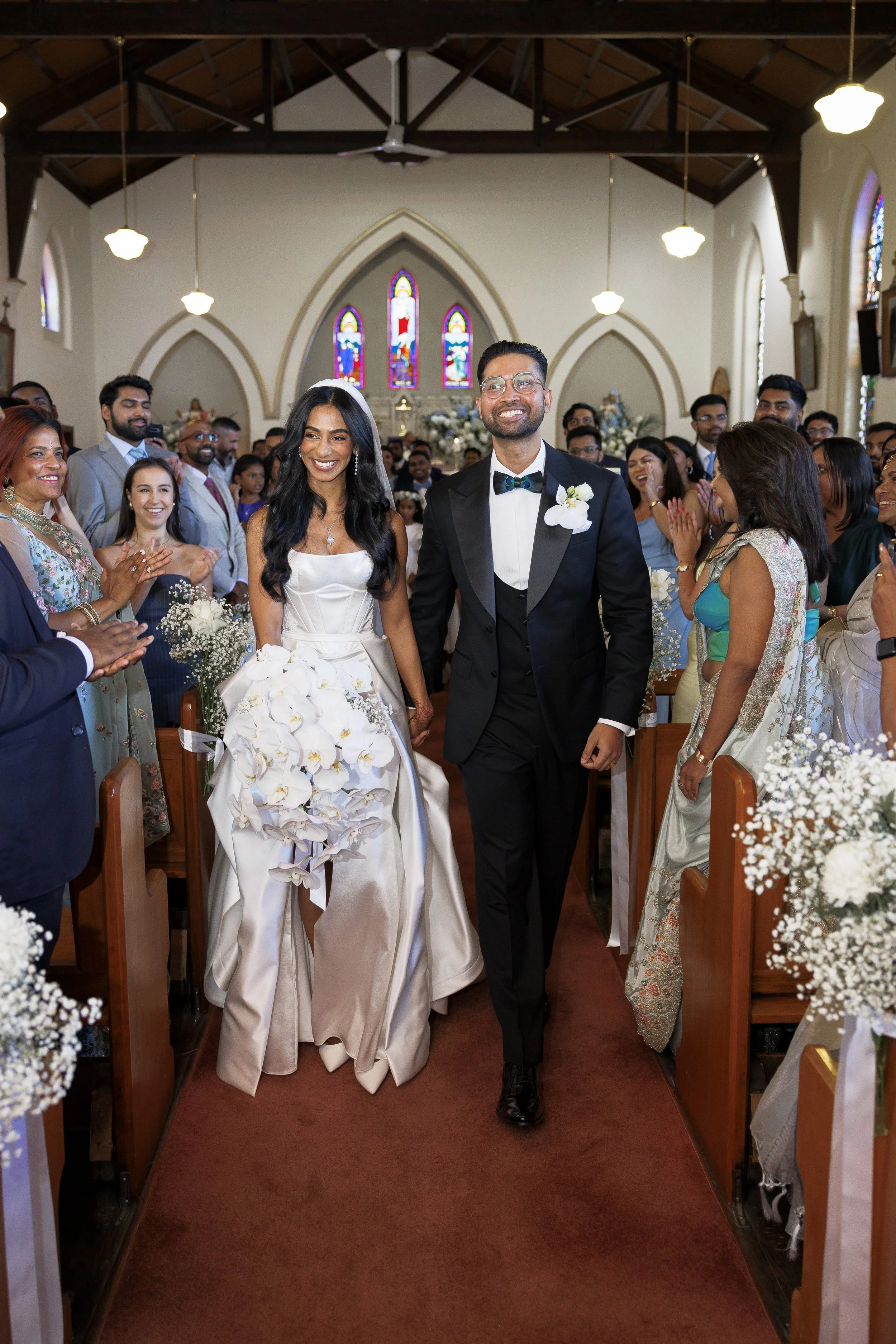 A bride and groom smiling and holding hands as they walk down the aisle of a church, surrounded by clapping guests, with stained glass windows and floral decorations visible in the background.