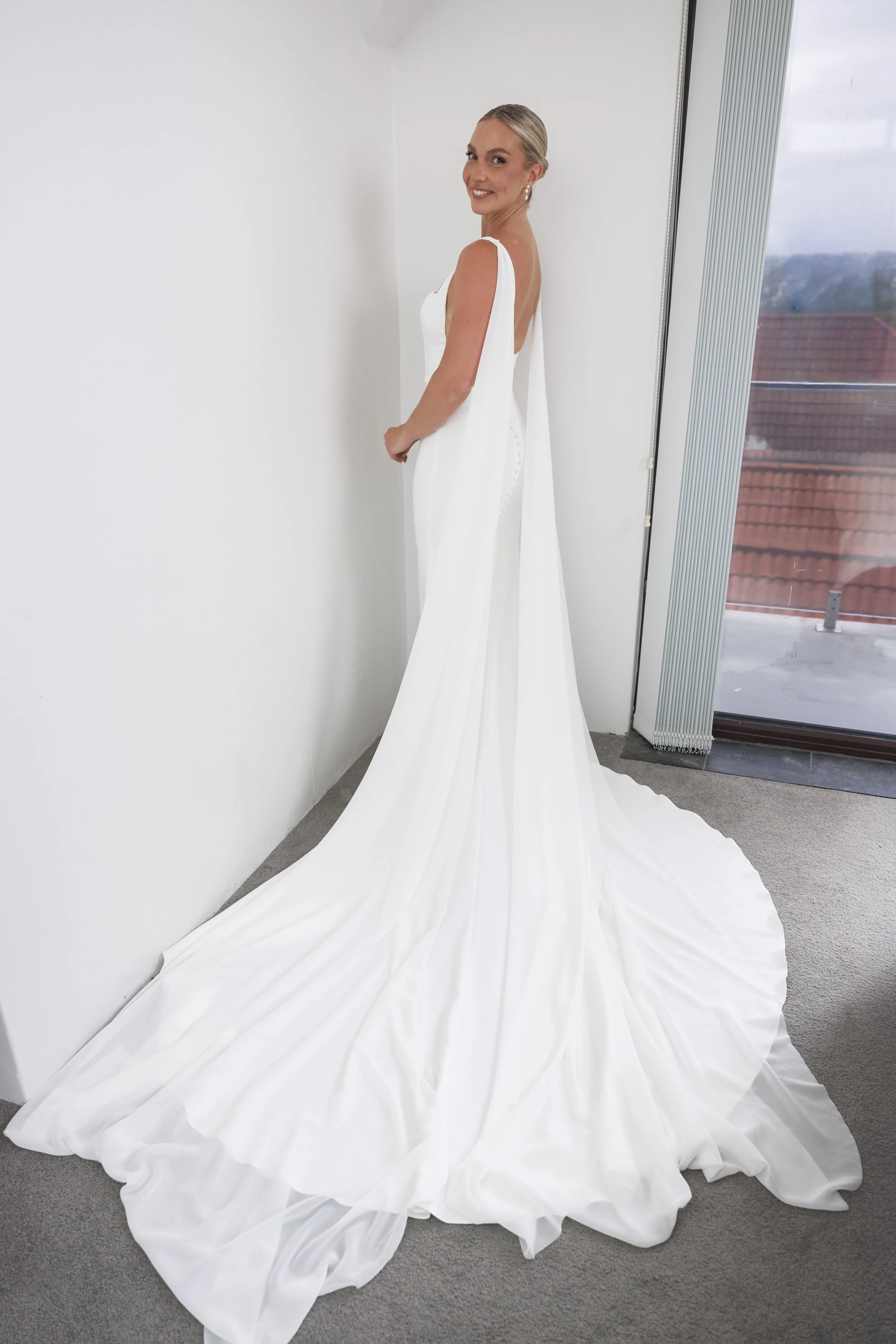 A woman in a white wedding dress with a long train is standing in a room near a window, smiling at the camera.