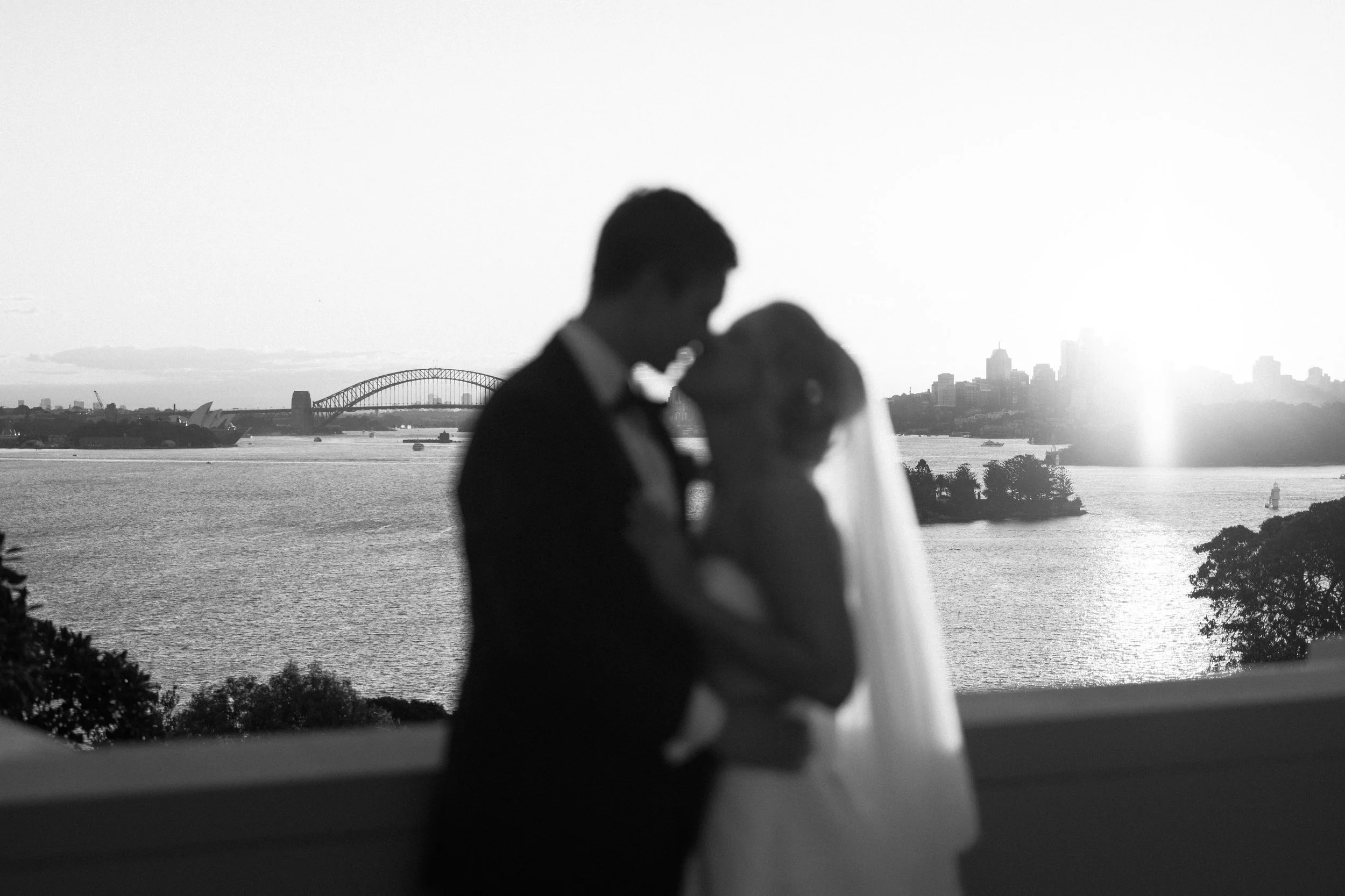 Silhouetted bride and groom kissing during sunset with Sydney Harbour Bridge and city skyline in background.