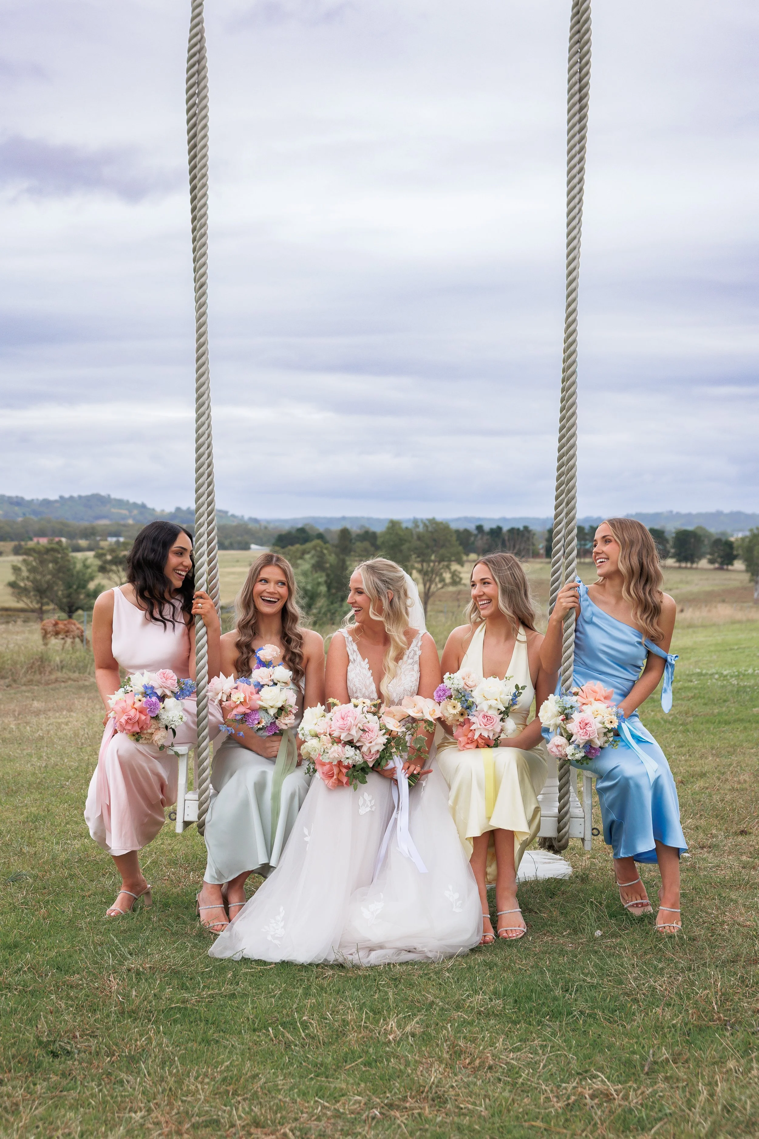 Group of five women, one in a wedding dress, sitting on a swing outdoors, holding bouquets of flowers, smiling and laughing.