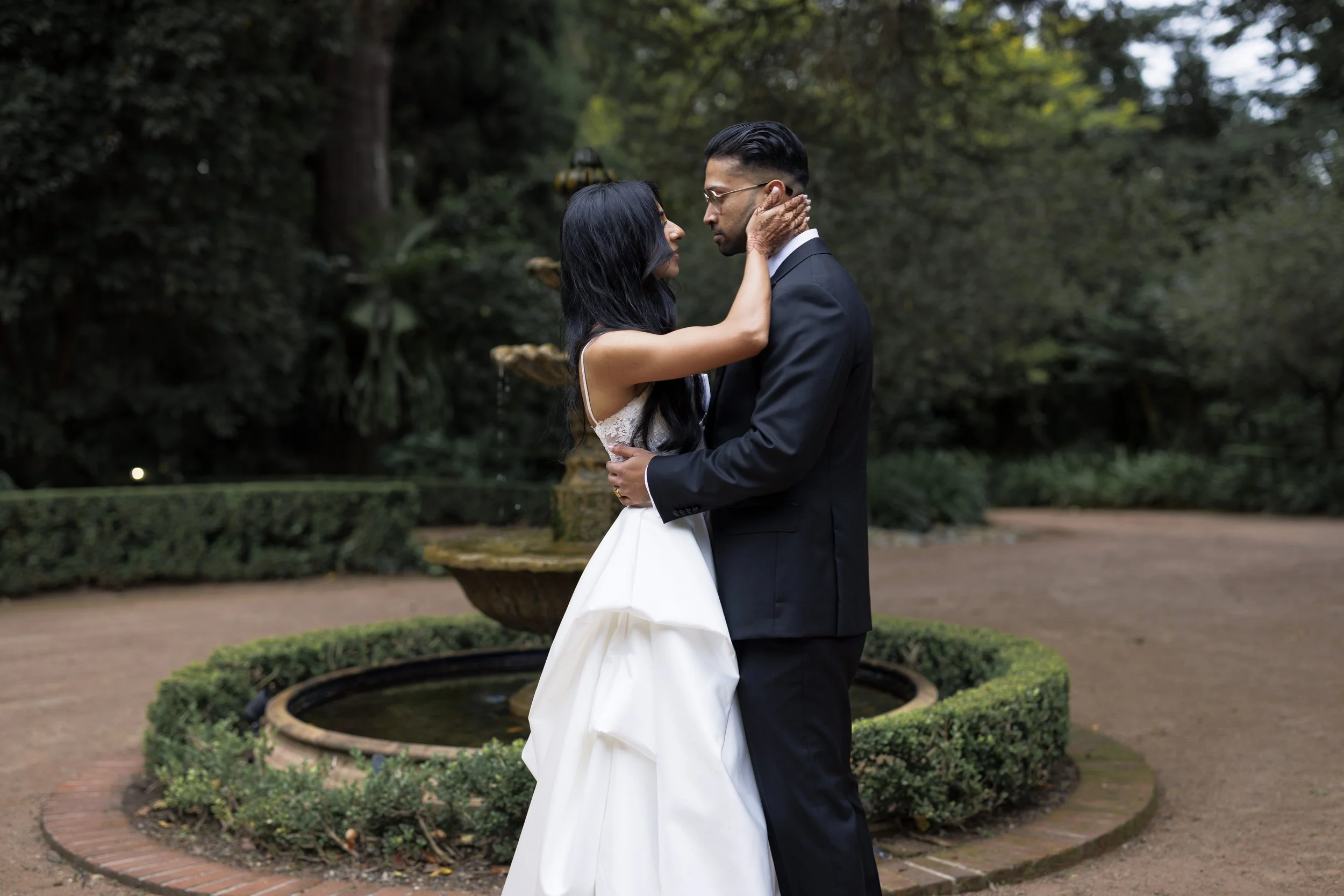 A bride and groom embrace in front of a fountain in a garden.