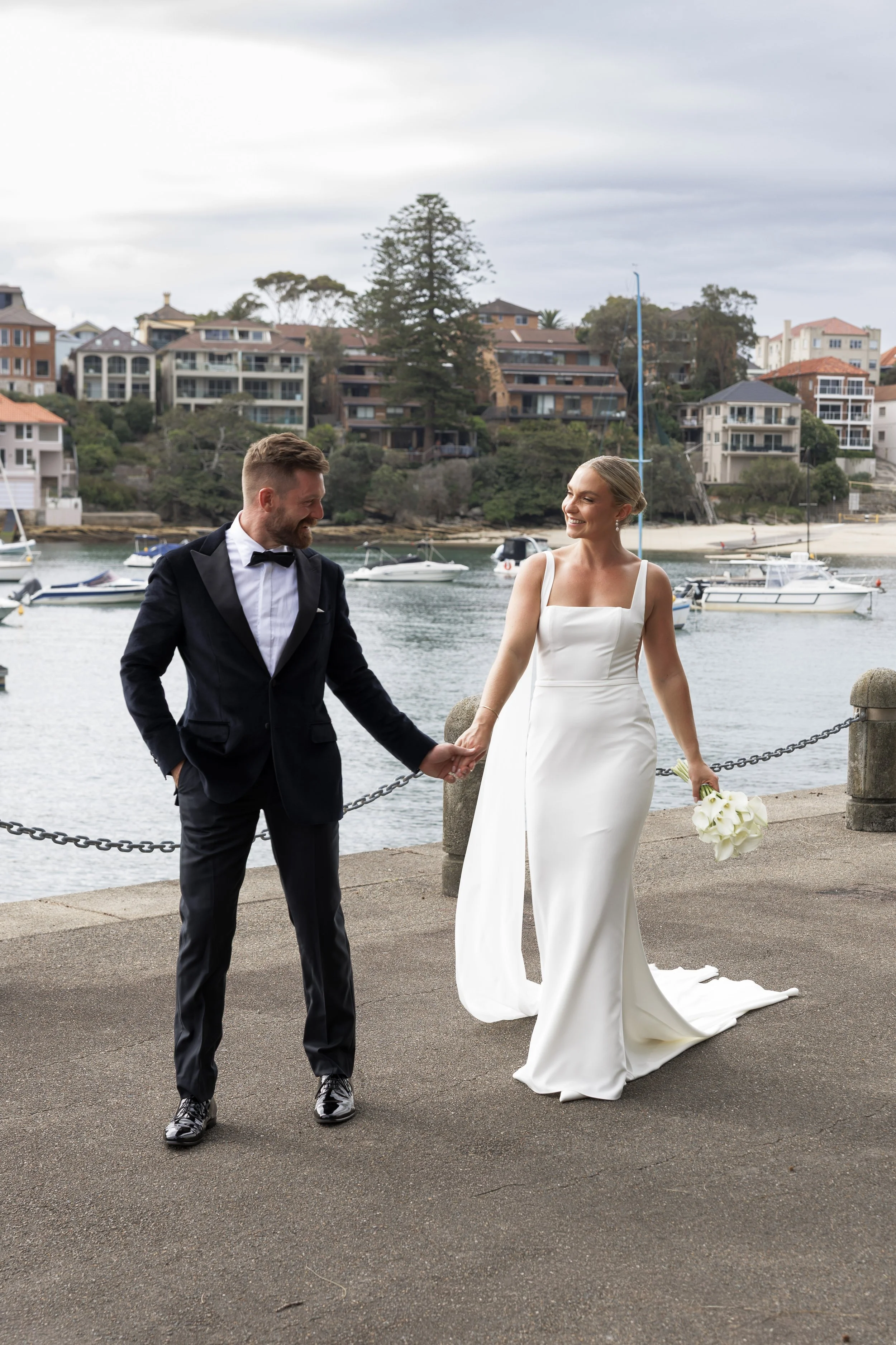A bride and groom holding hands on a waterfront promenade with boats and houses in the background.