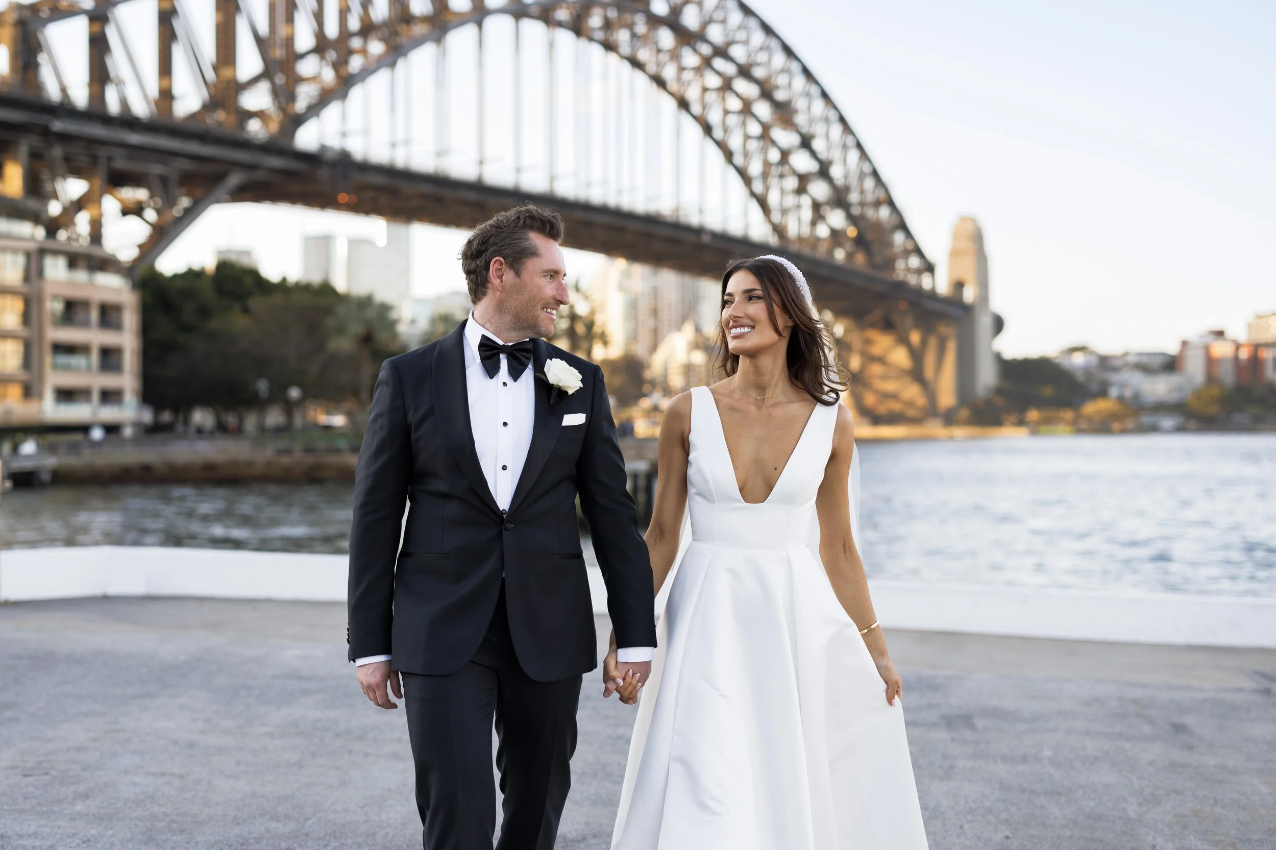 A newlywed couple walking hand in hand near a river with a large steel bridge and city buildings in the background during sunset.