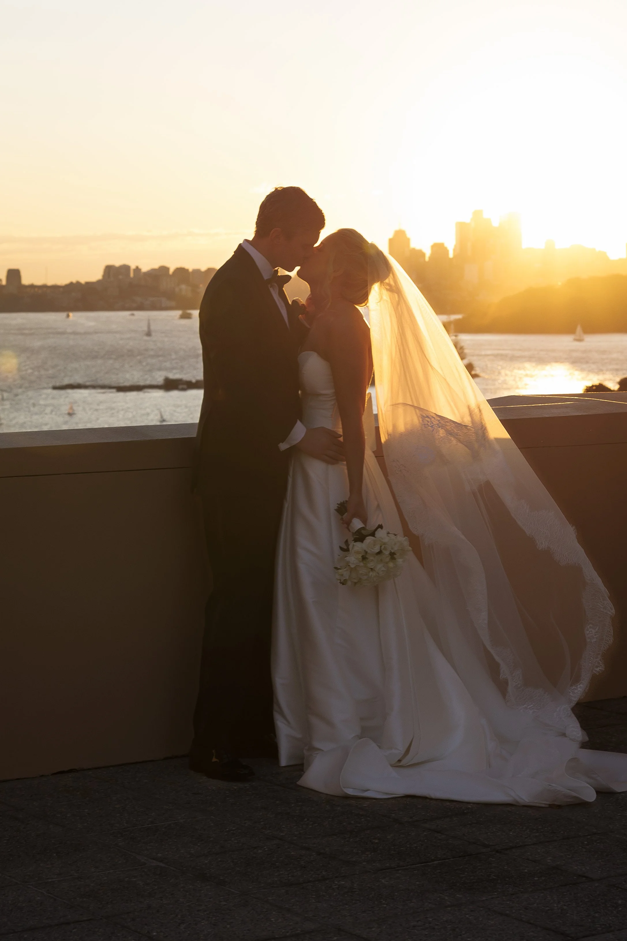 A bride and groom sharing a kiss at sunset near a body of water, with a city skyline in the background.