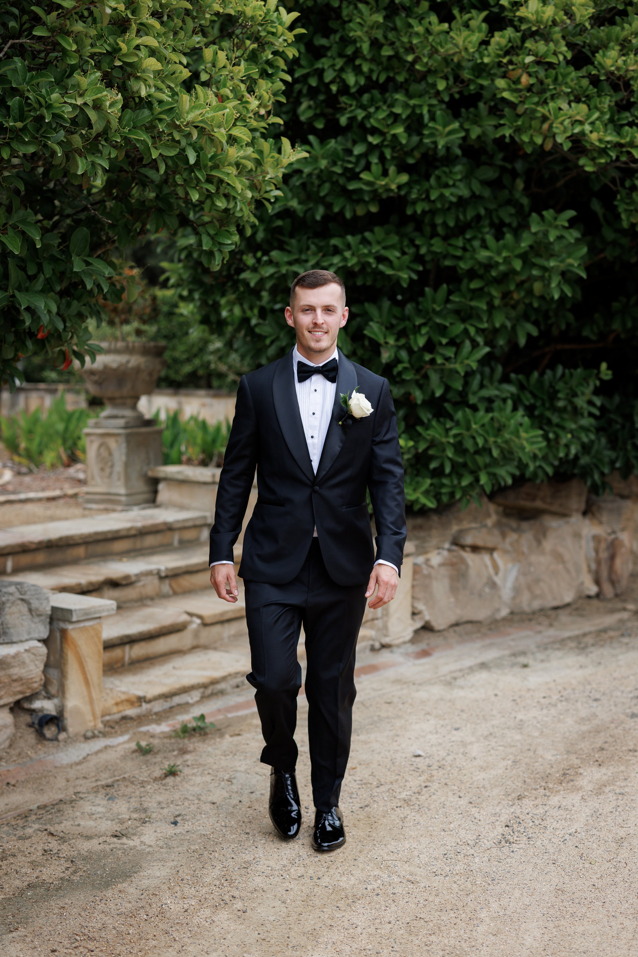 A man in a tuxedo with a bow tie and a white boutonniere walking outdoors on a dirt path, with greenery and stone steps in the background.