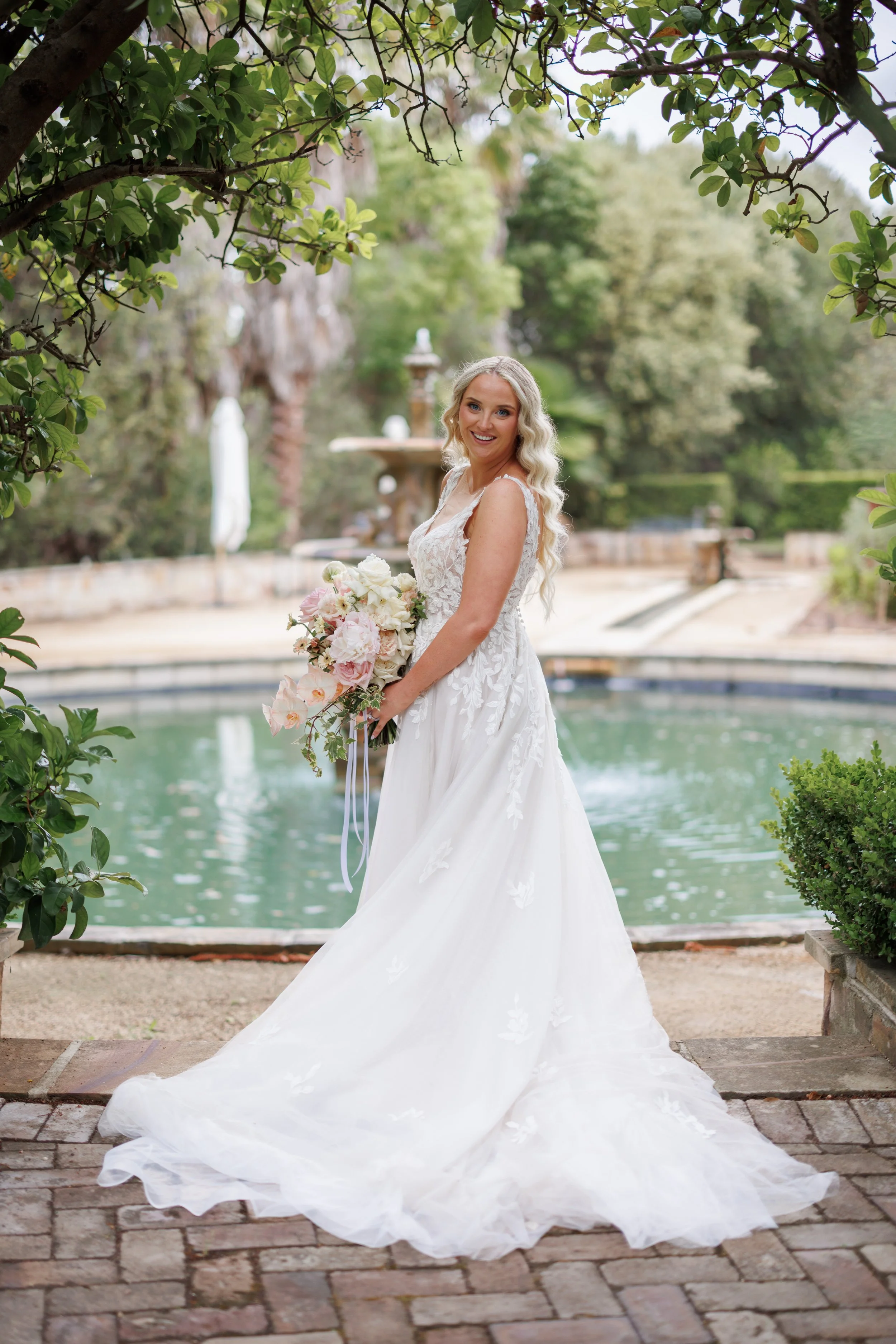 A bride in a white wedding dress holding a bouquet, standing outdoors near a pond, surrounded by trees and greenery.