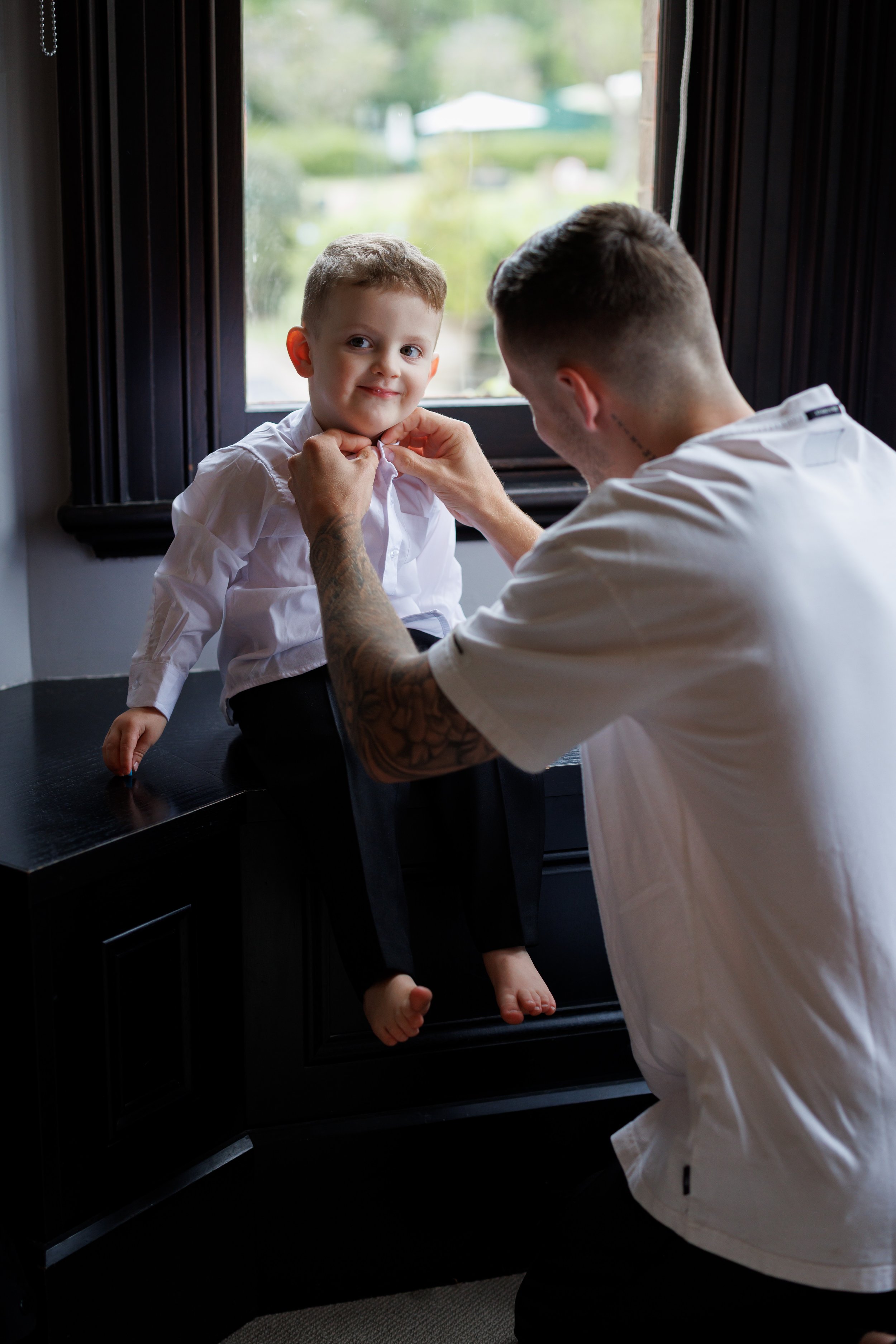 A man is helping a young boy with a collar, sitting by a window with greenery outside, inside a home.