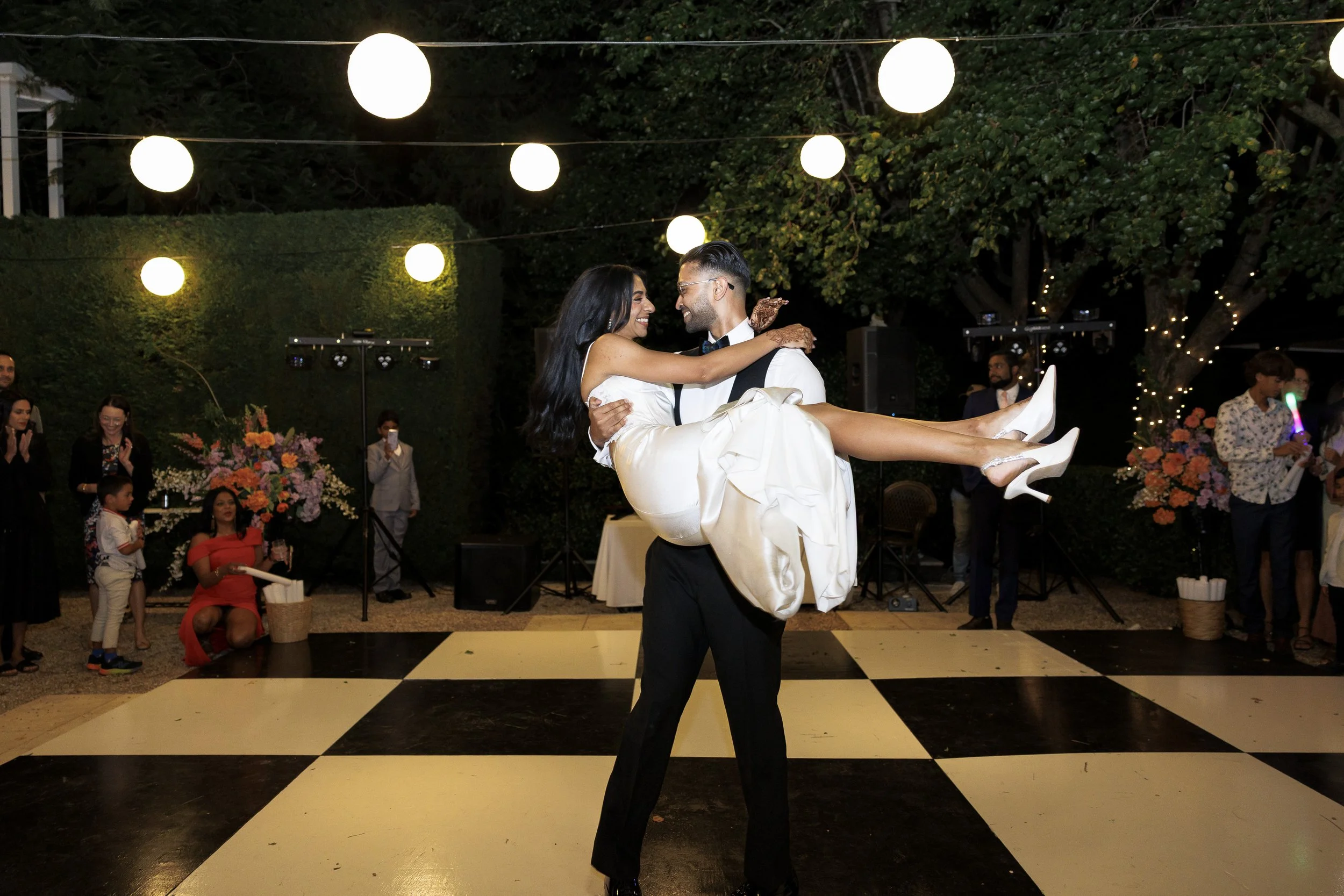 A man in a tuxedo lifts a woman in a white dress during a dance at an outdoor wedding reception, with string lights hanging overhead and guests watching.