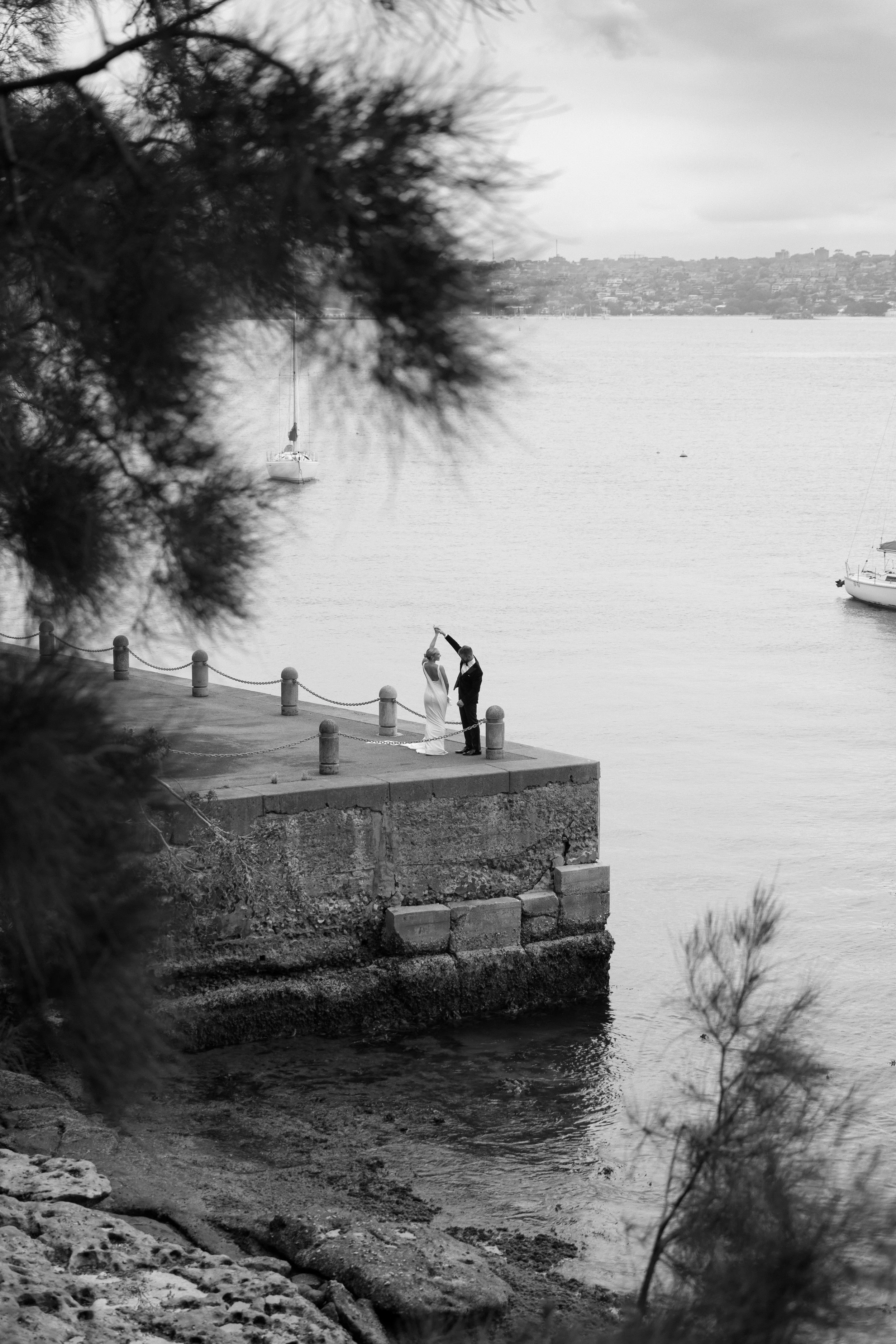A black and white photograph of a couple on a dock, with the man dipping the woman during their wedding or engagement photo. They are surrounded by water with sailboats in the background, and trees framing the scene.