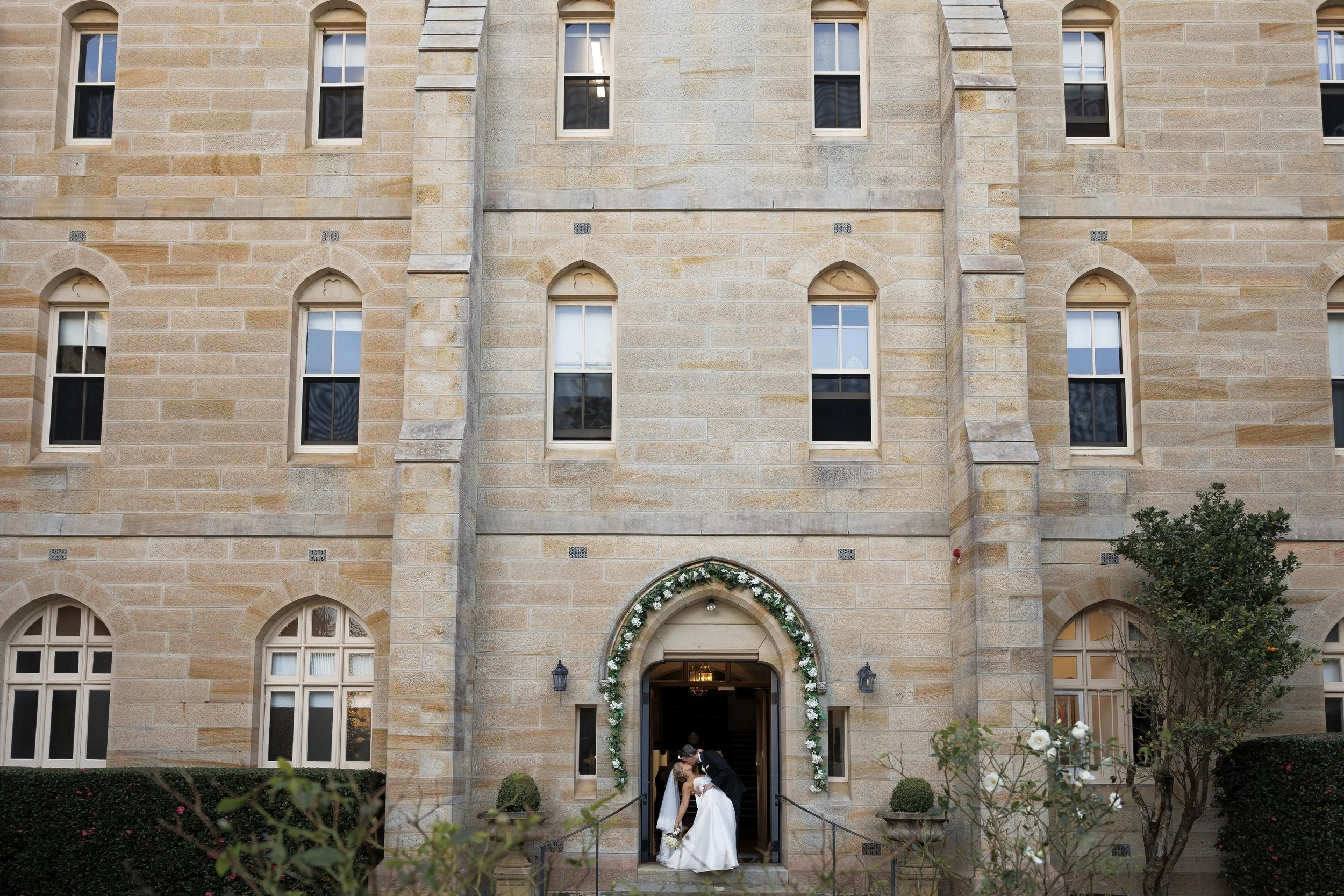 Bride and groom kissing at the entrance of a stone building decorated with flowers
