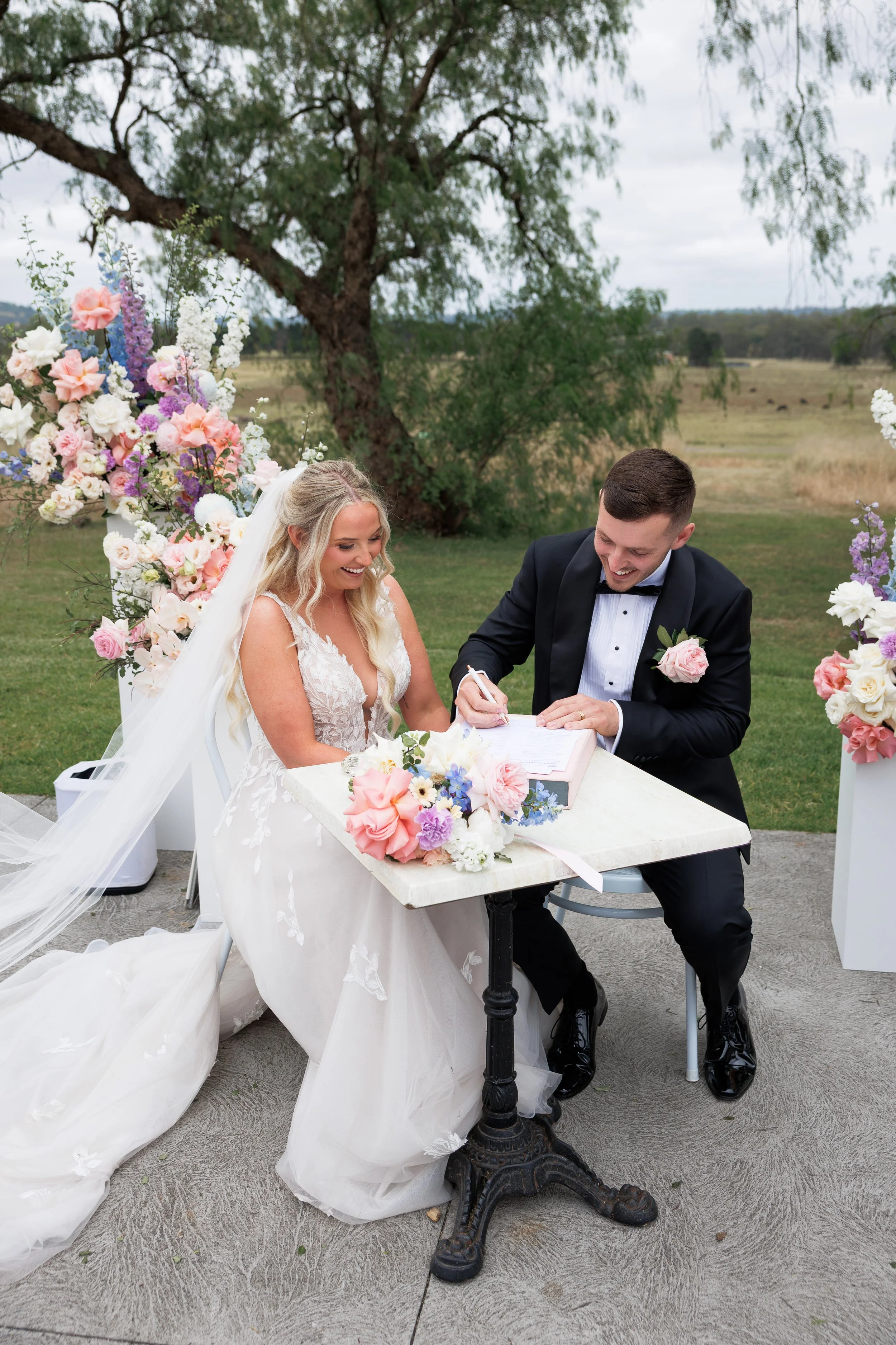 A bride and groom sit at a table outdoors, signing a marriage document, surrounded by pastel-colored flowers, with a large tree and open field in the background.