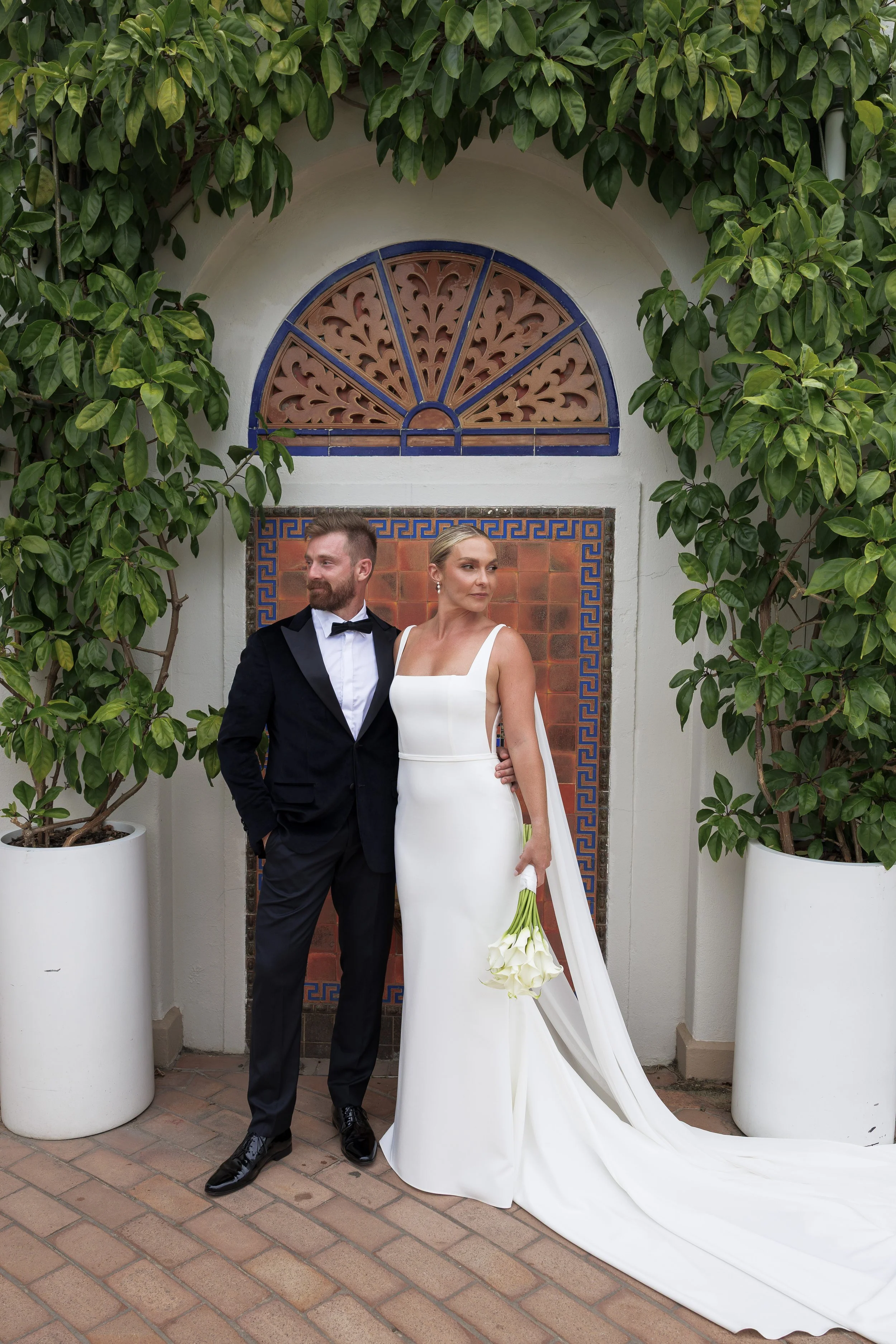 A bride and groom in formal wedding attire standing outside in front of a decorative wall with tiles and greenery, with the bride holding a bouquet of white calla lilies.