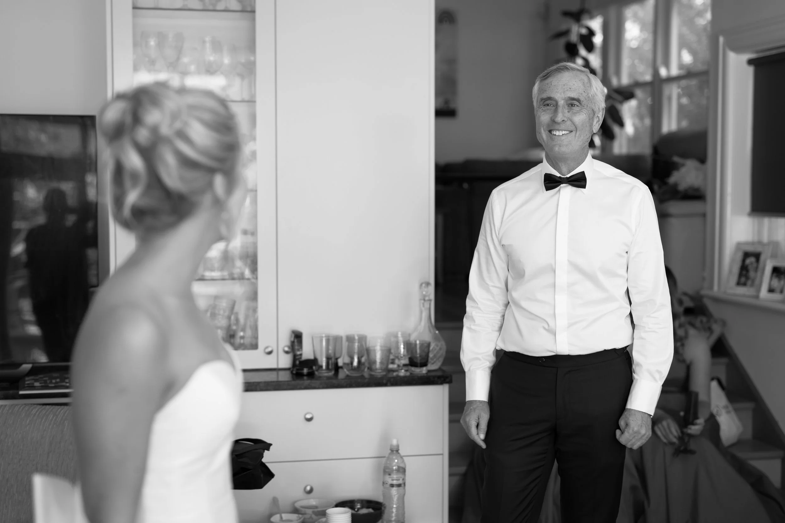 A woman in a wedding dress looking at a man in formal attire with a black bow tie in a room with a window and decorative items.