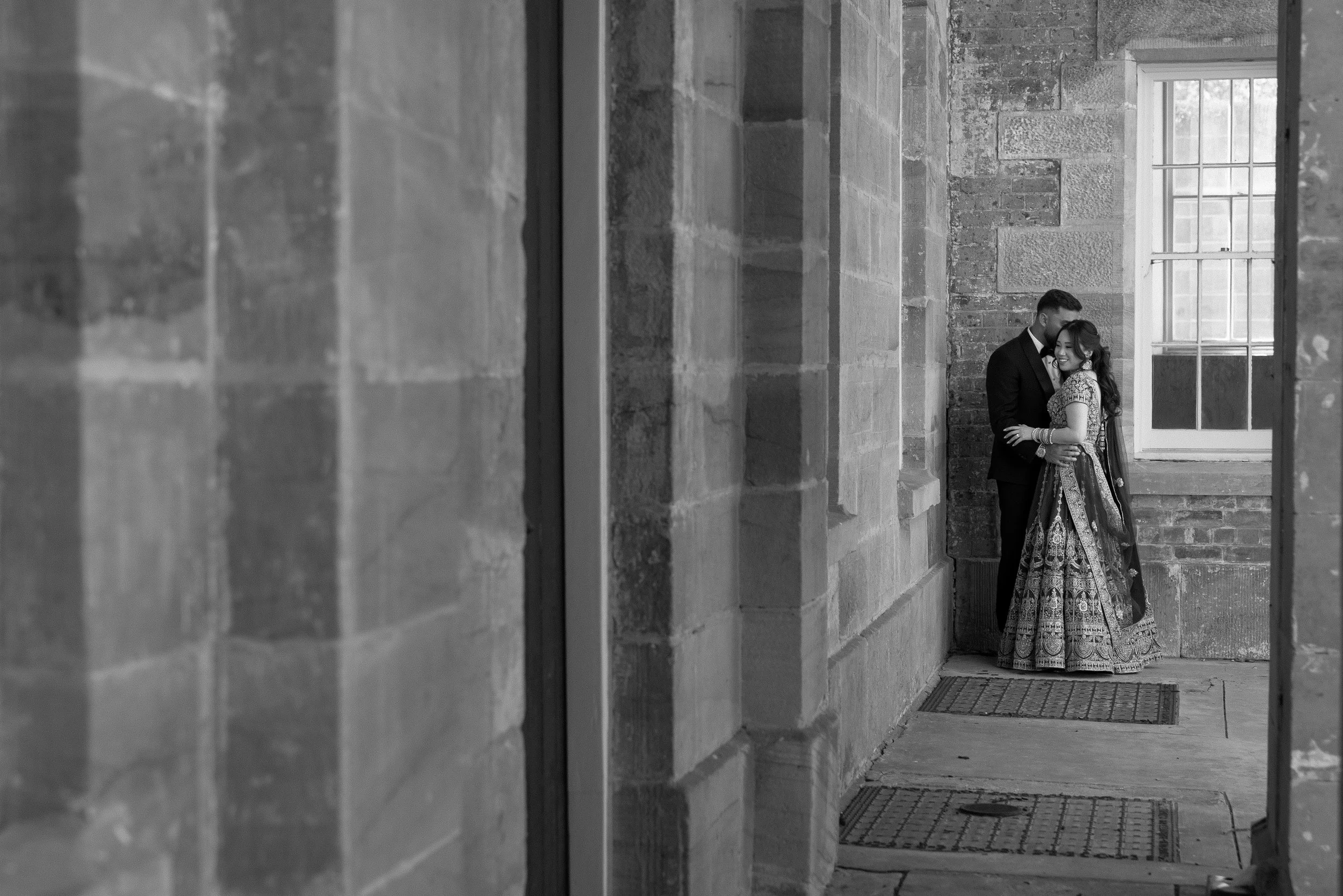 A couple dressed in formal attire standing close together, smiling, in an industrial-style space with brick walls and large windows