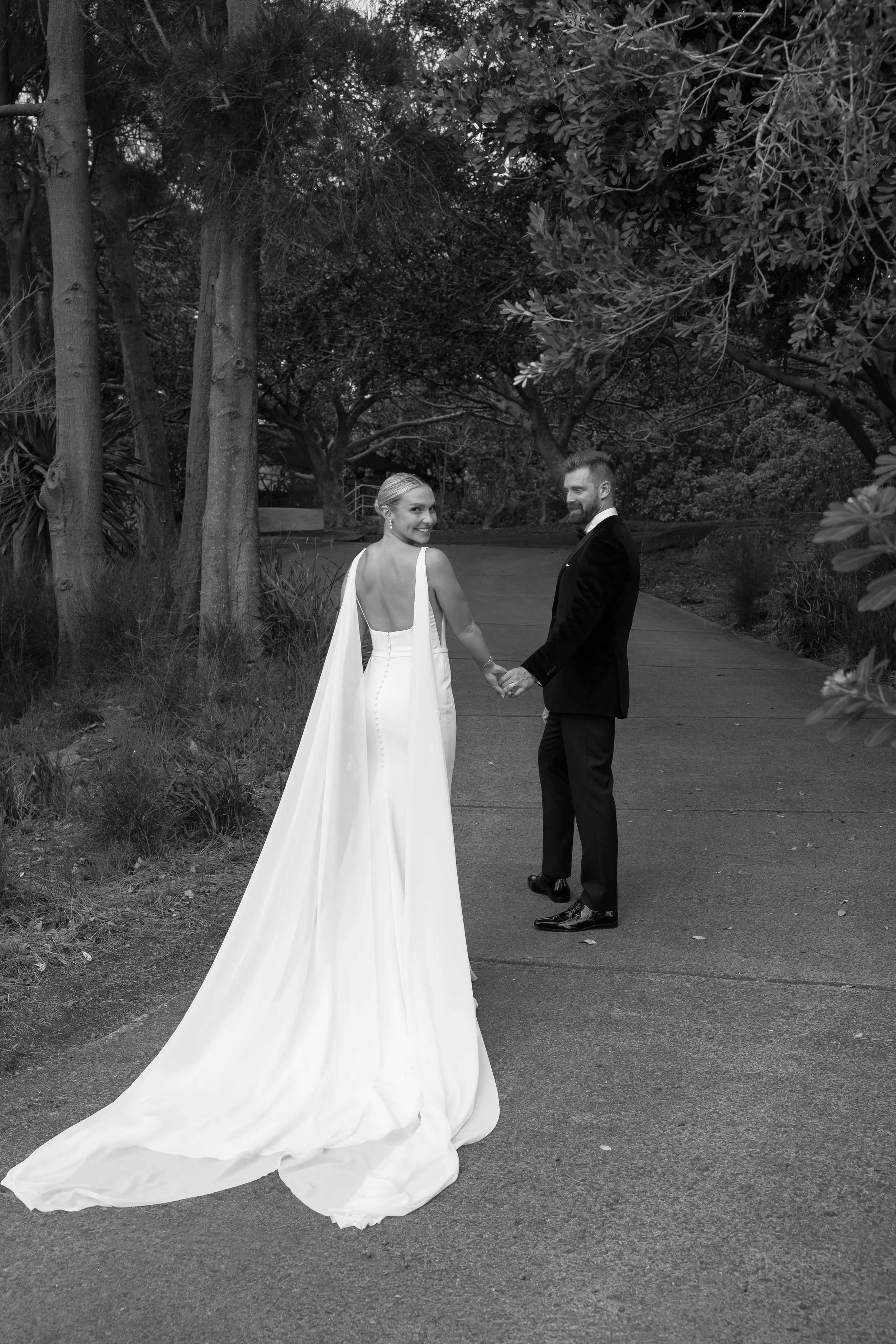 Black and white photo of a bride and groom holding hands and walking on a paved path through a wooded area. The bride is wearing a long white gown with a cape, and the groom is dressed in a dark suit. They are smiling and looking back toward the came