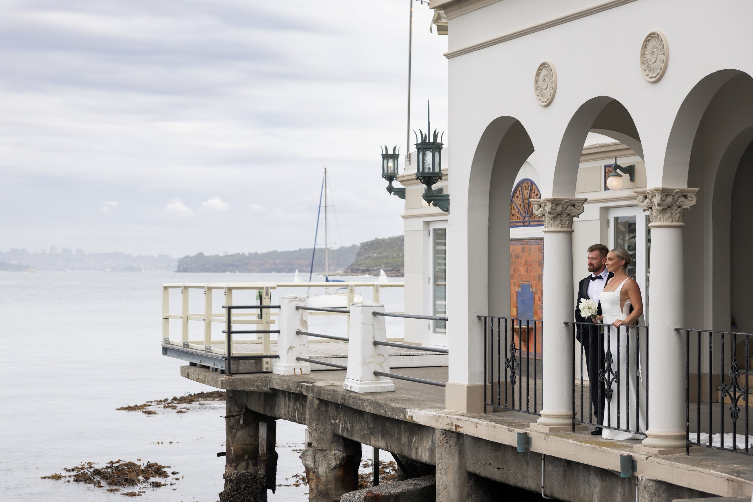 A newlywed couple standing on a balcony overlooking the water, with sailboats in the distance. The bride is holding a bouquet and wearing a white wedding dress, while the groom is in a tuxedo.
