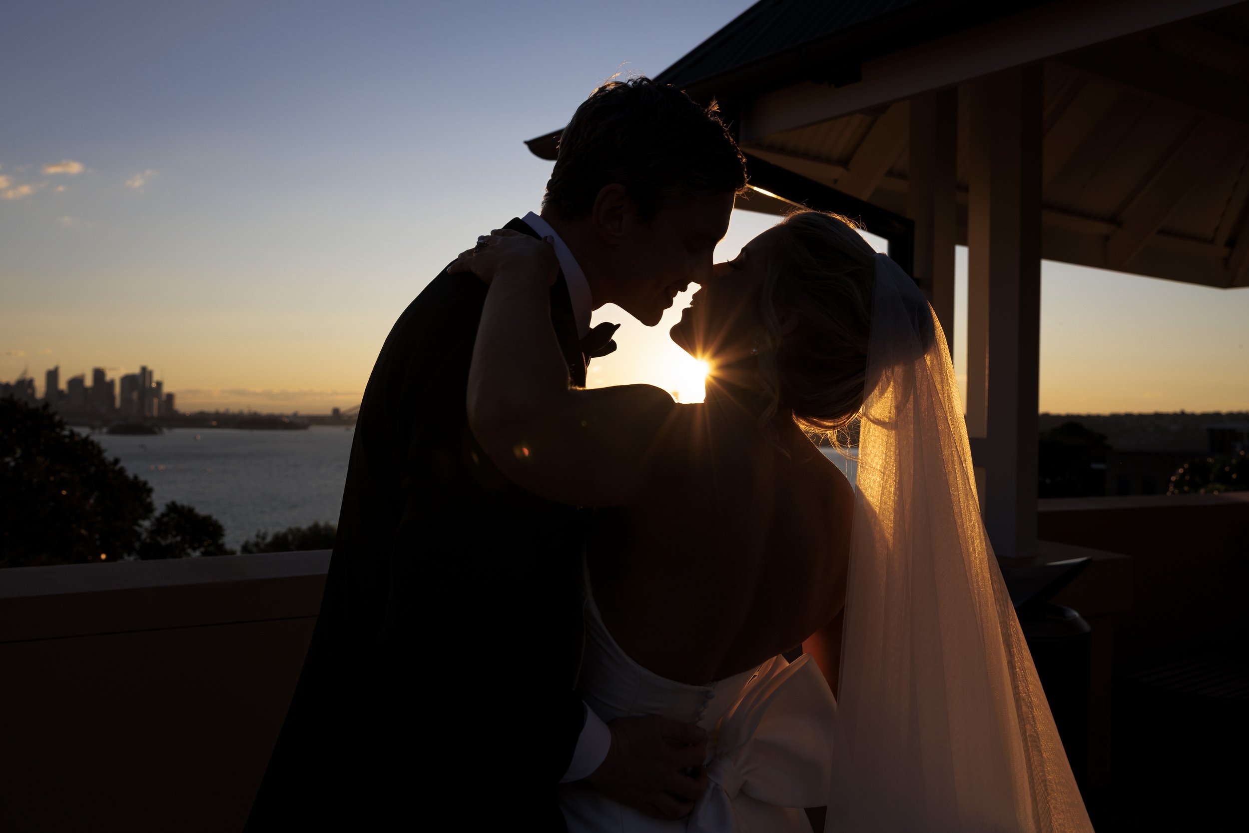 Silhouette of a couple at sunset, with the man leaning in towards the woman who is wearing a veil, on a balcony overlooking water and city skyline.