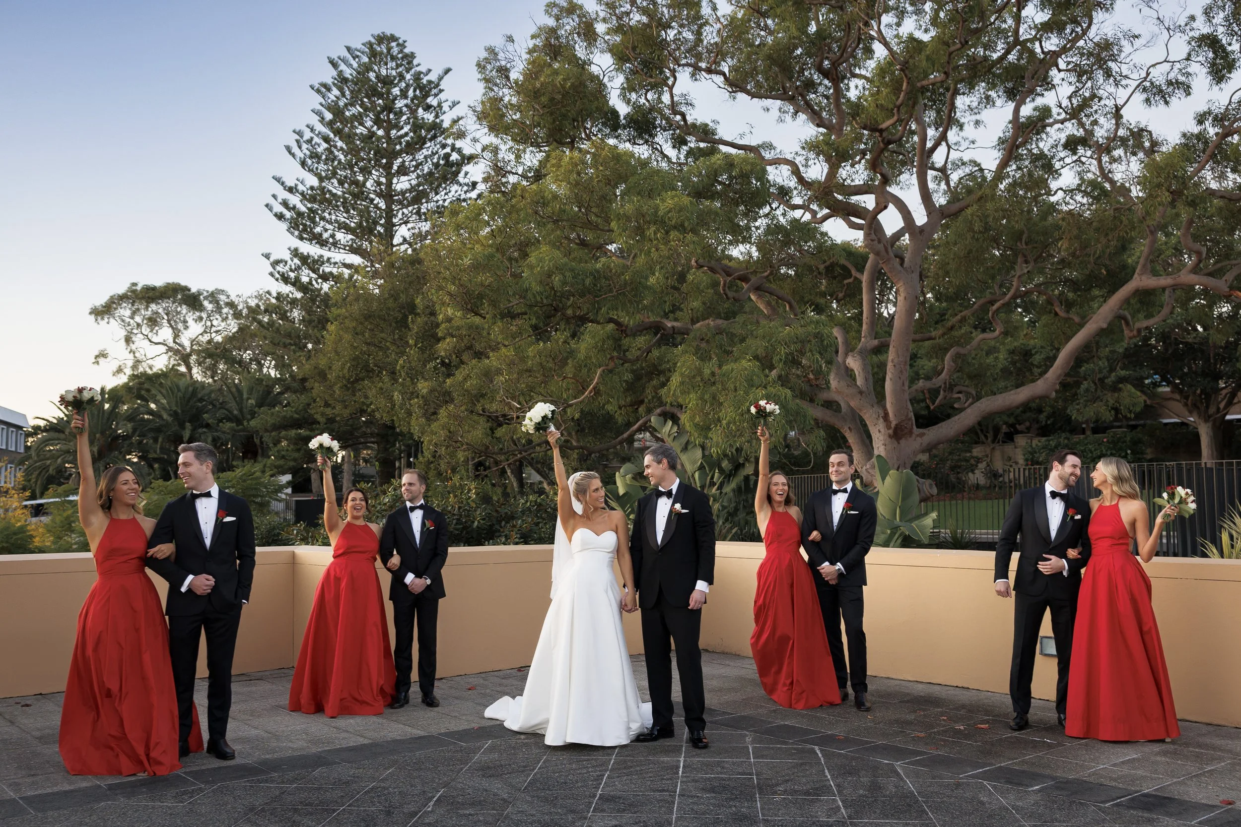 Wedding party standing outdoors near a large tree, with the bride and groom in the center holding hands, surrounded by bridesmaids in red dresses and groomsmen in black tuxedos. Some are holding bouquets and raising them in celebration.