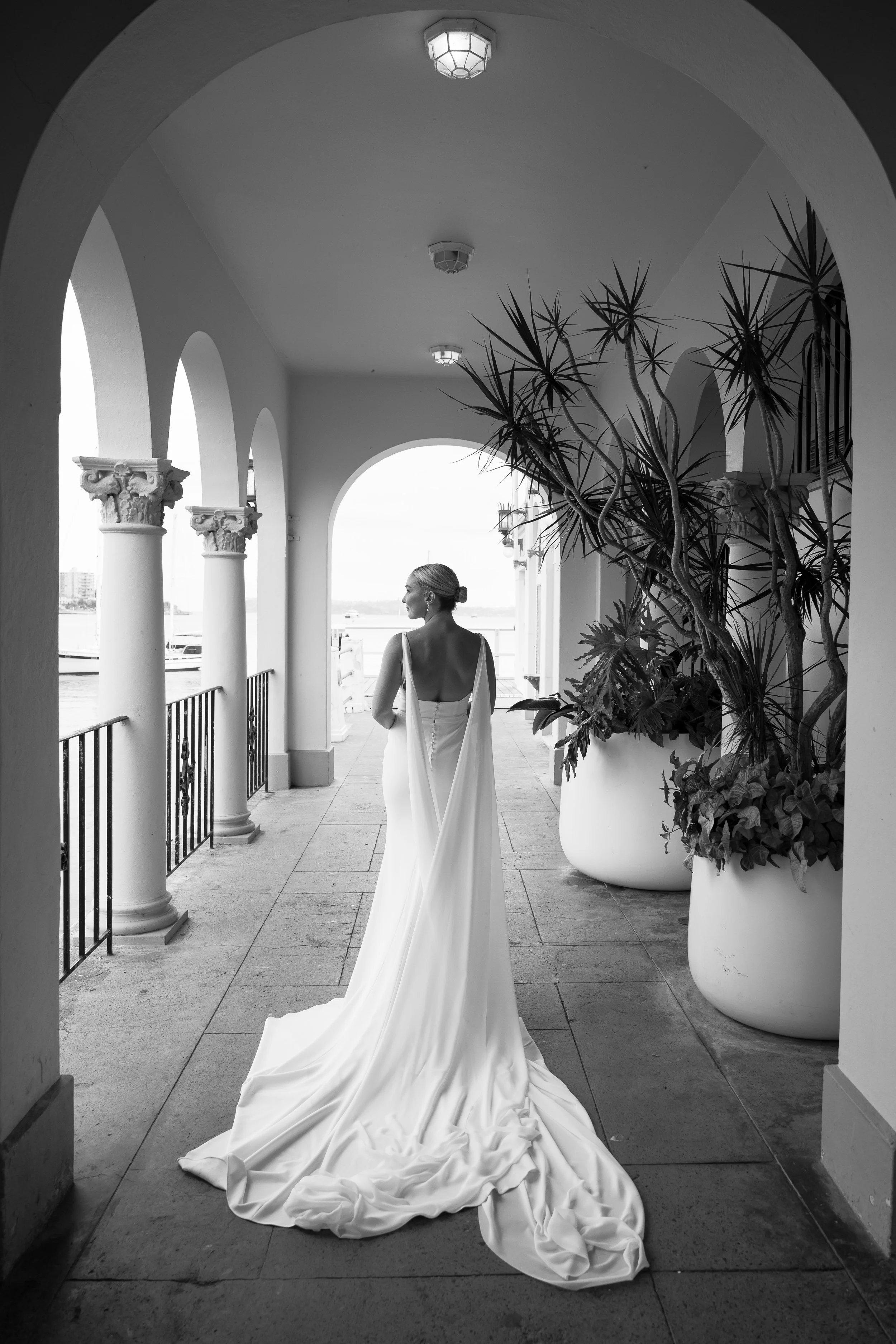 A woman in a white wedding dress stands on a balcony with classical columns, overlooking a body of water, in black and white.