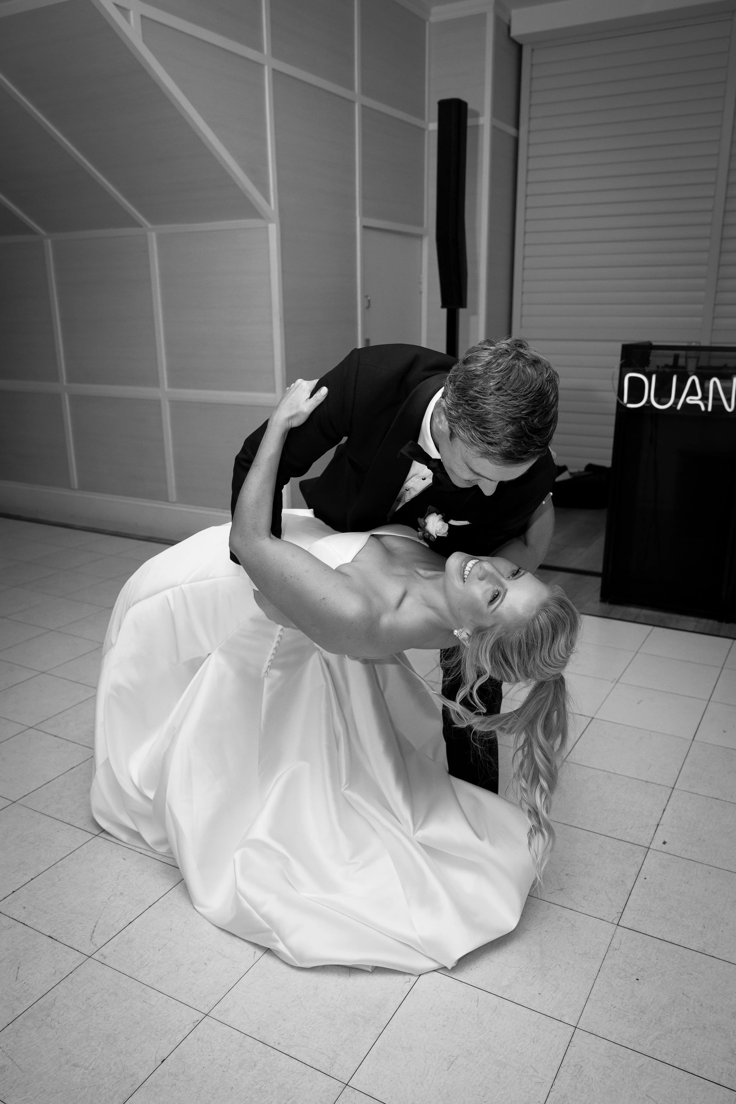 A bride and groom sharing a dance at their wedding reception, with the groom dipping the bride who is smiling and looking at the camera in a black-and-white photo.