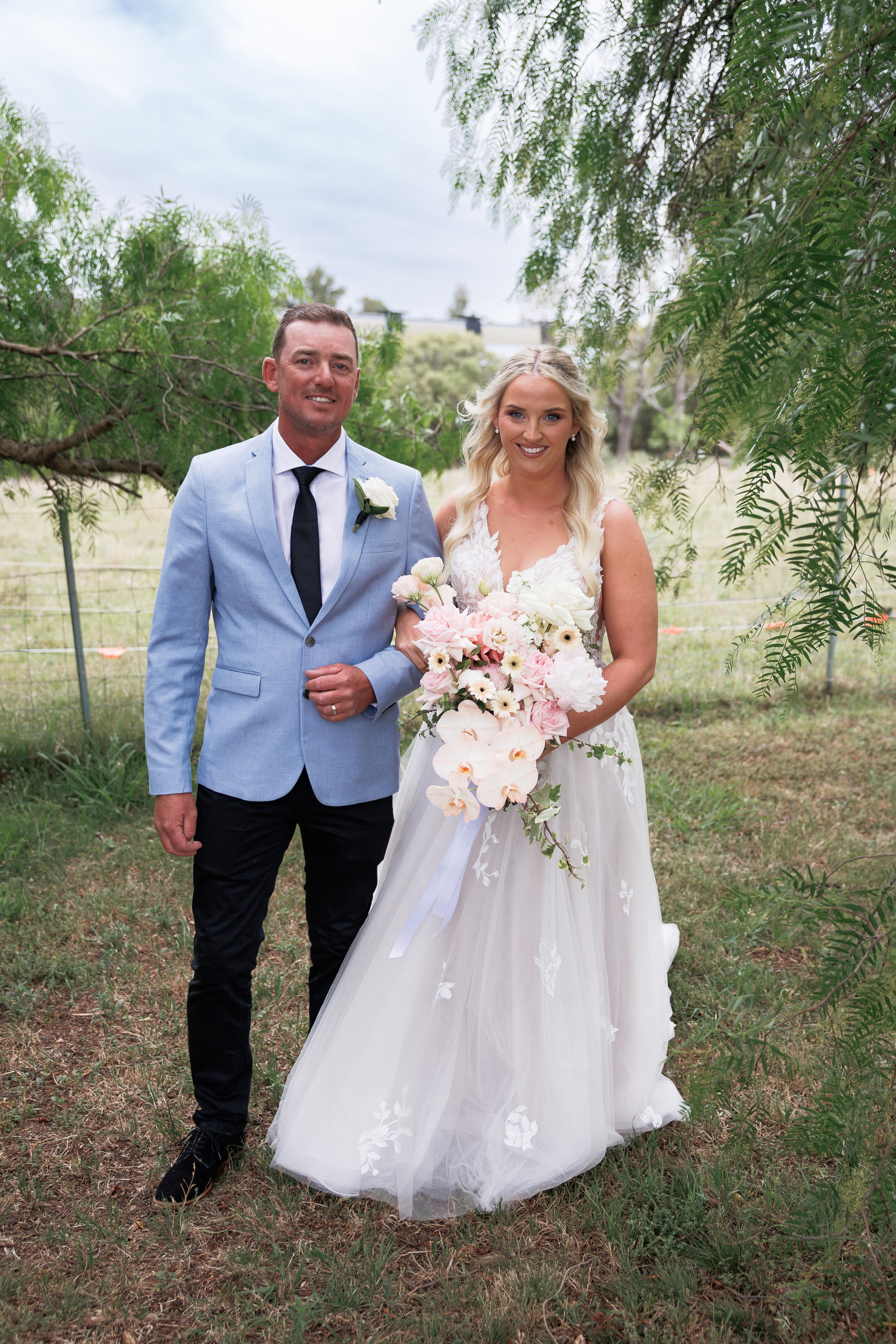 A bride and a man, possibly her father, standing outdoors on a grassy area with trees and a fence in the background. The bride is holding a large bouquet of pink and white flowers and wearing a white wedding dress, while the man is wearing a light bl